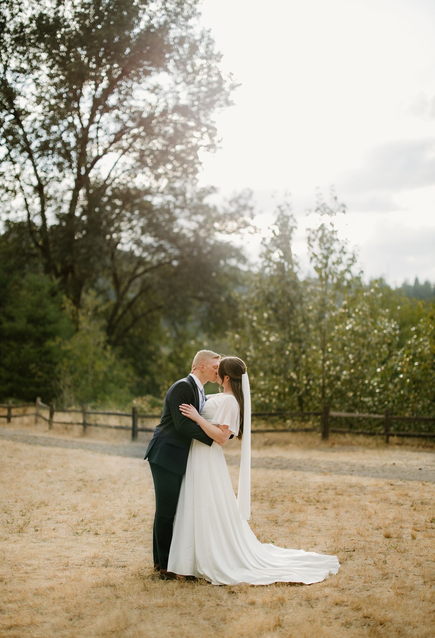 Bride and groom share a romantic kiss at golden hour in a scenic outdoor wedding venue with trees.
