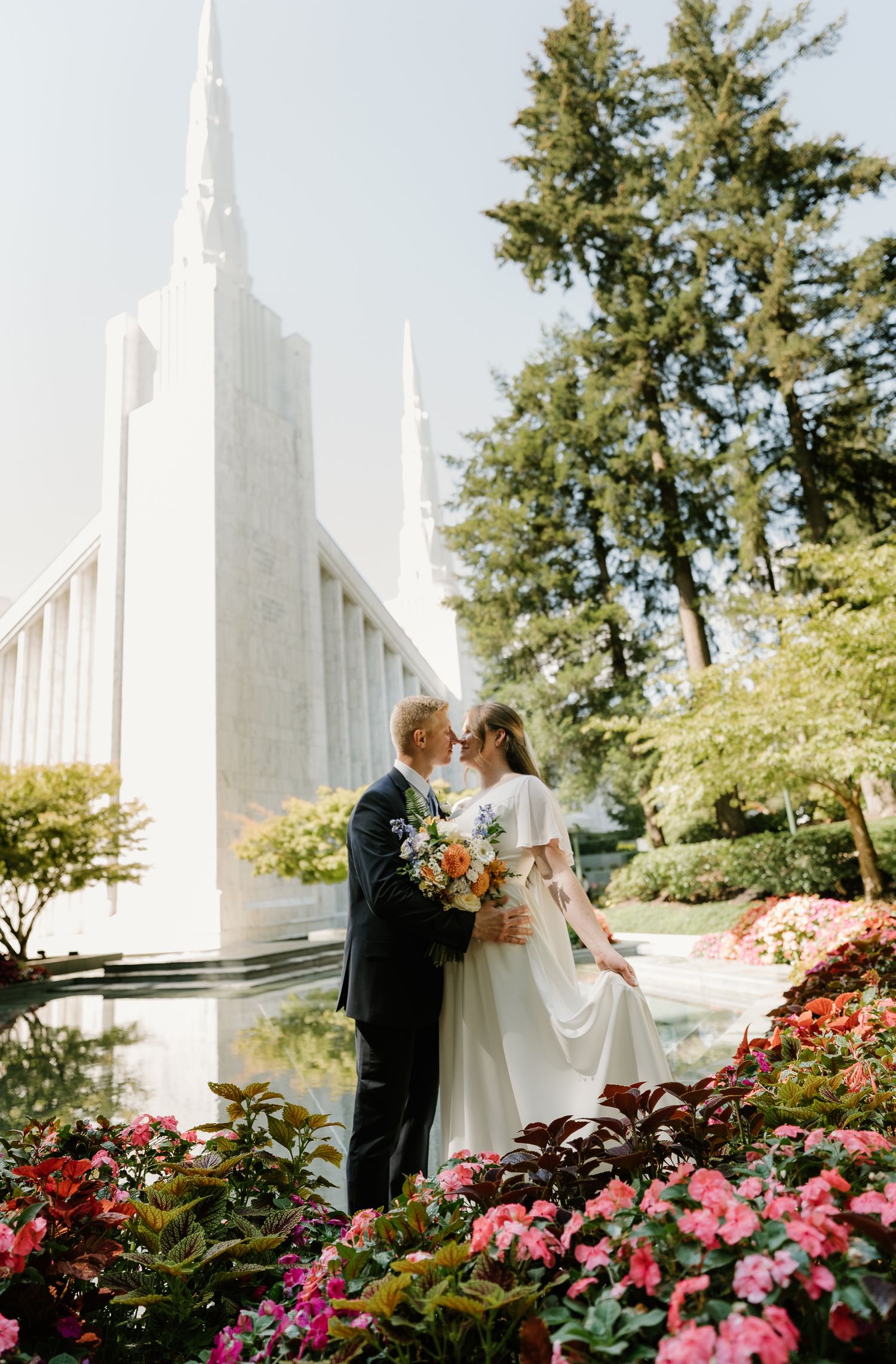 Bride and groom share a kiss among colorful flowers outside a white LDS temple.