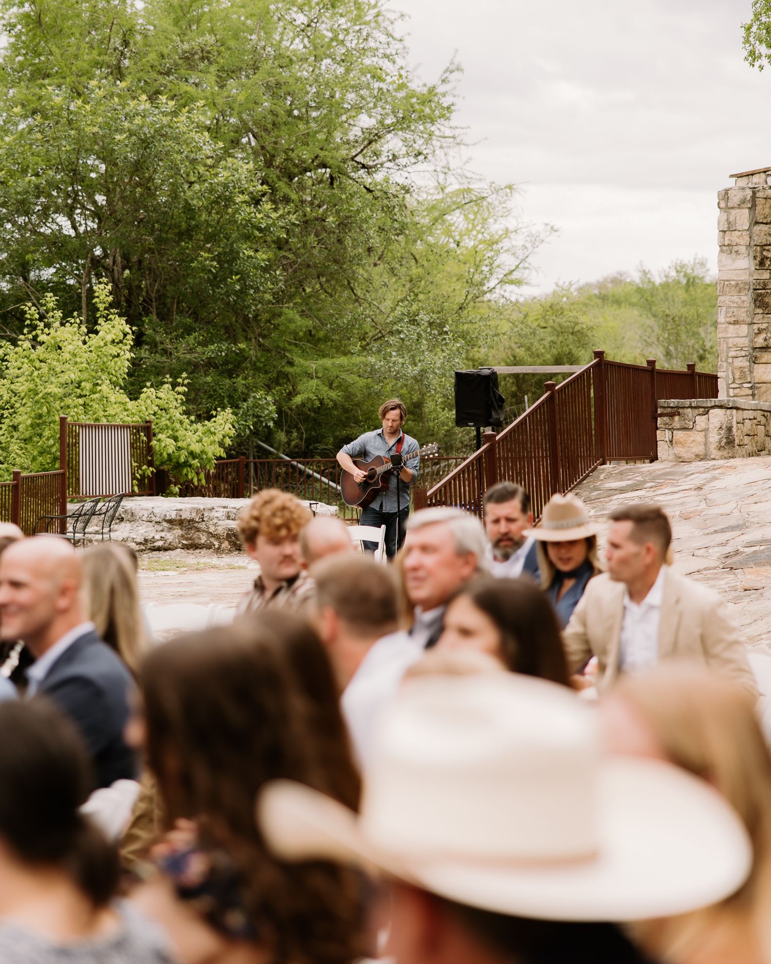 Live acoustic guitarist performs for seated wedding guests outdoors at a rustic stone venue.
