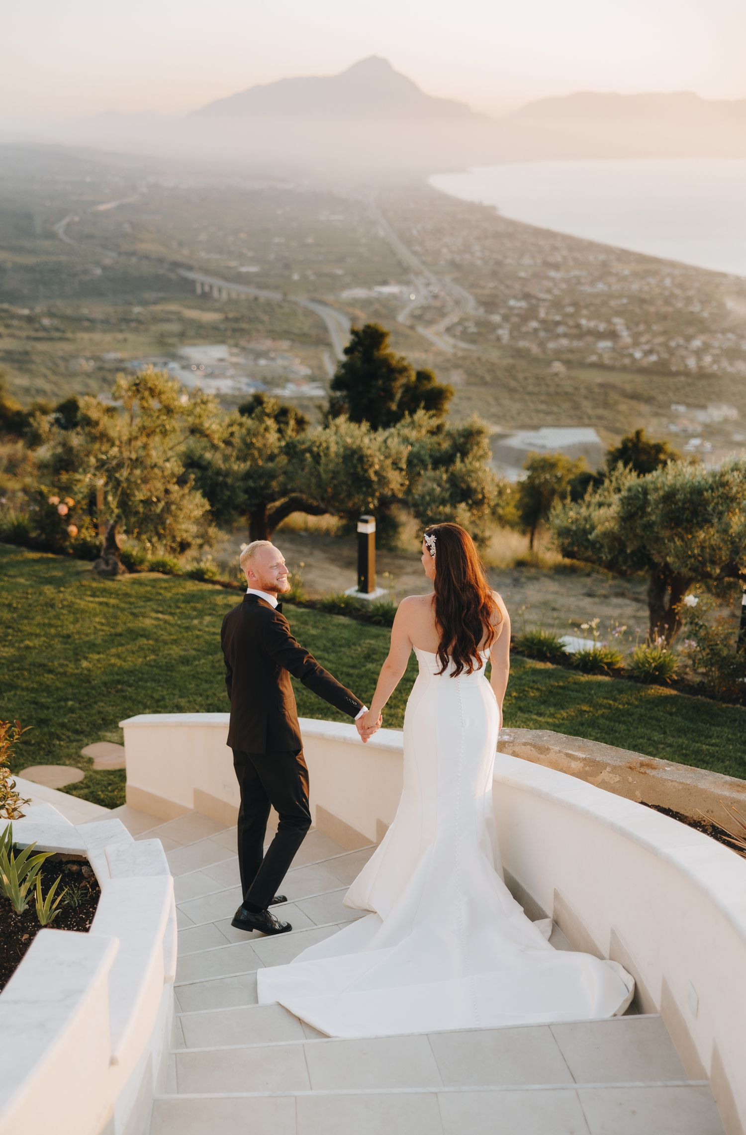 Bride and groom holding hands on scenic terrace steps with Mediterranean coastal view at golden hour.