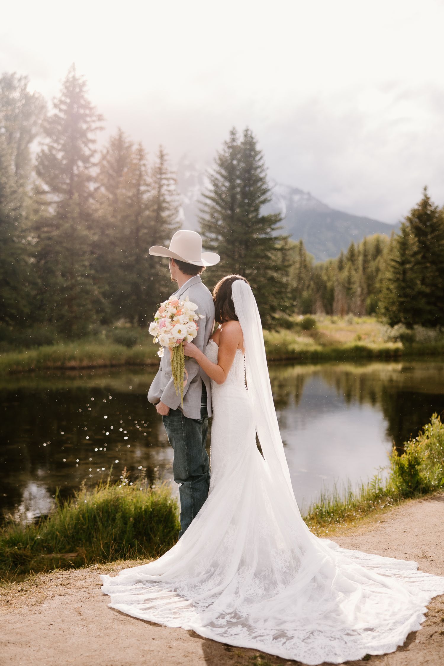 Bride and groom stand by a mountain lake, groom in cowboy hat, bride in lace gown with long train.
