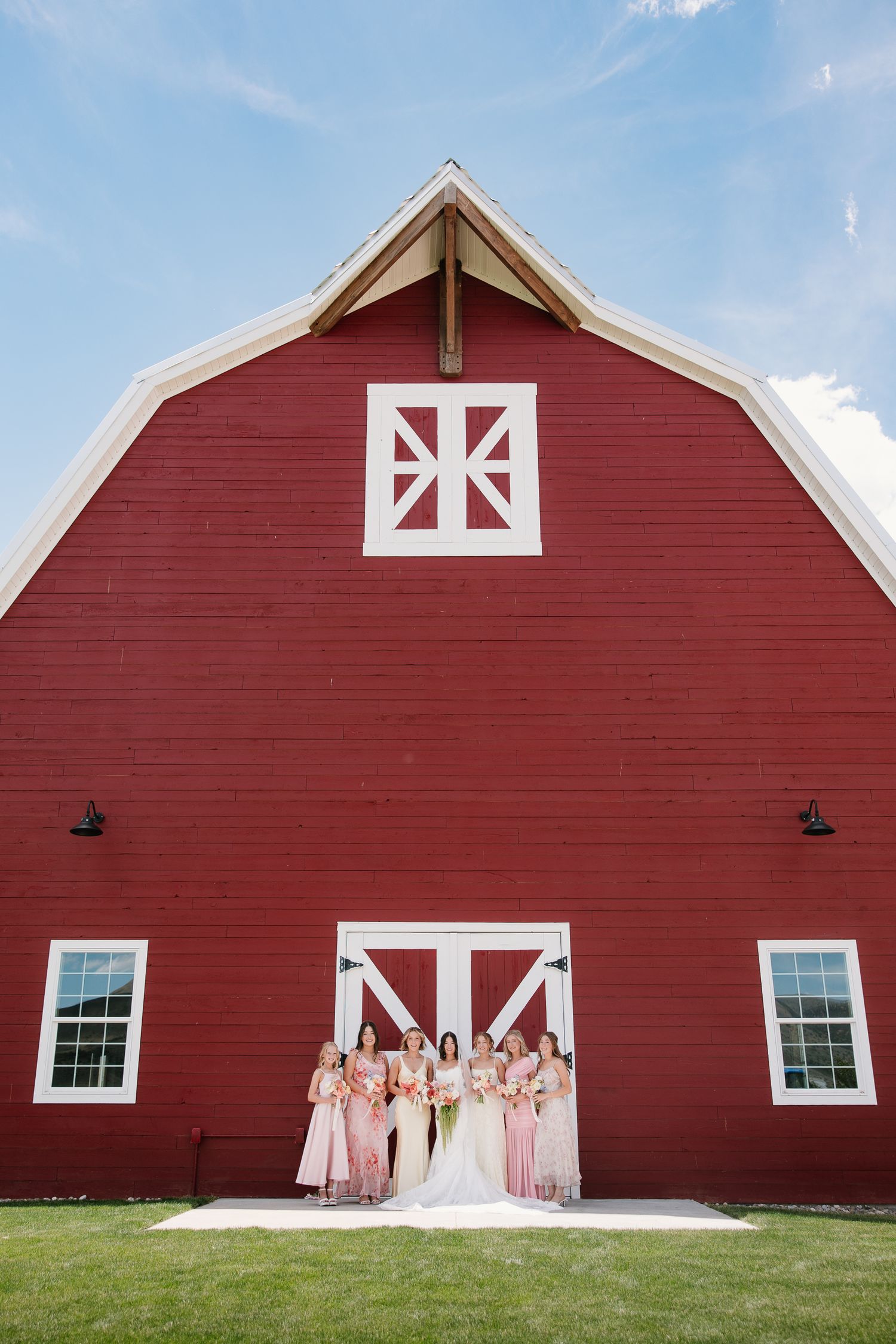 Bride and bridesmaids in pink dresses posing in front of a classic red barn on a sunny wedding day.
