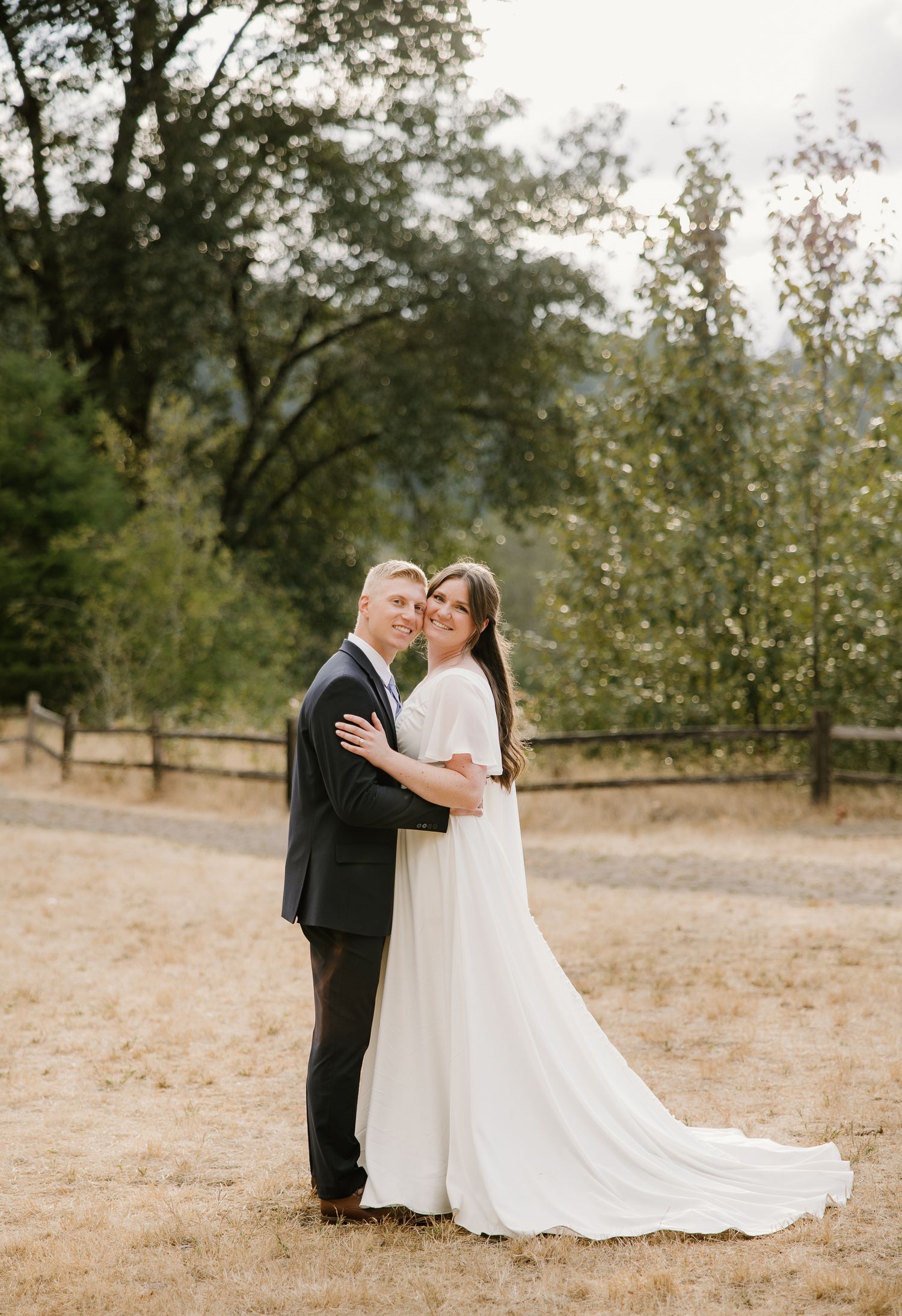 Bride in white gown and groom in dark suit embrace outdoors on golden field with trees and fence backdrop.