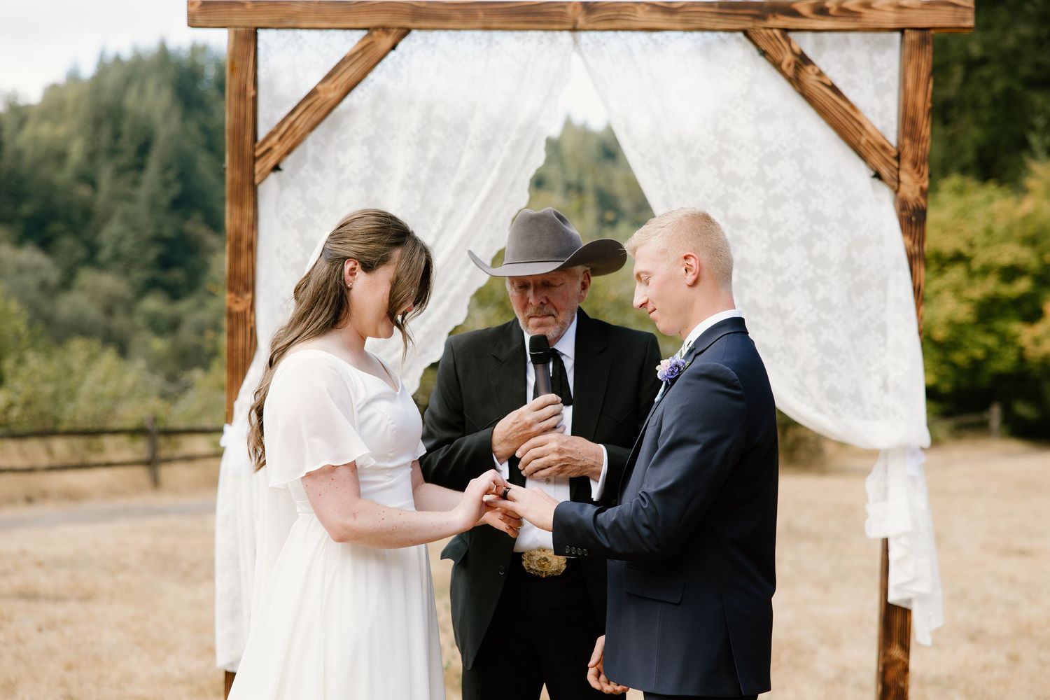 Officiant in hat leads ring exchange ceremony for couple under draped wooden wedding arch.