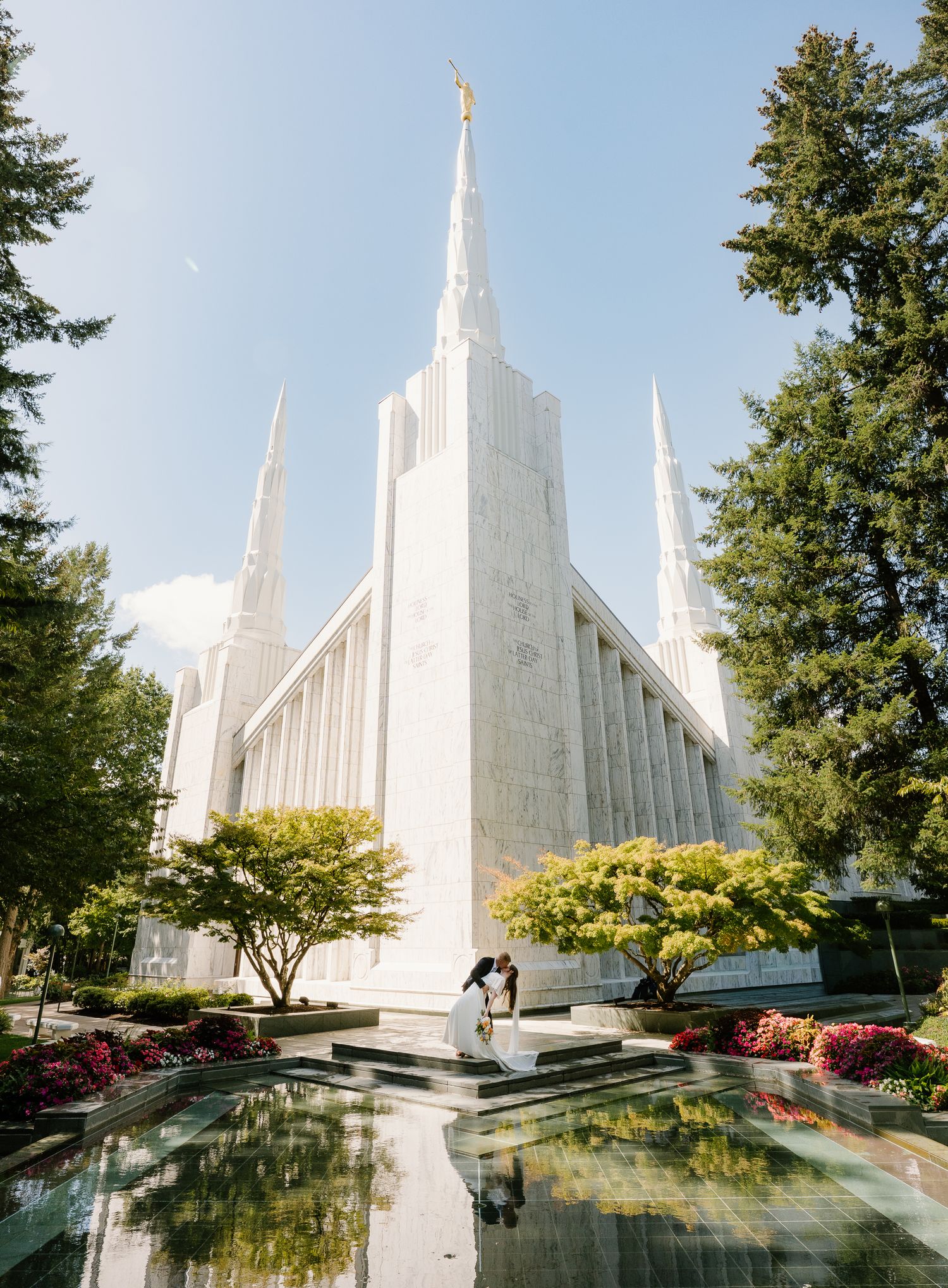 Bride and groom kiss near a reflecting pool in front of a white LDS temple spire.