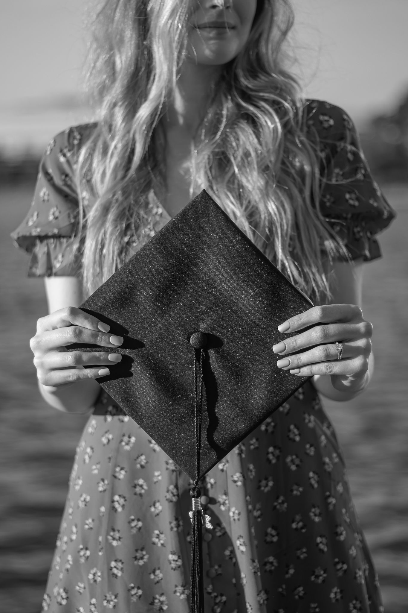 Black and white photo of woman holding graduation cap toward camera near water.
