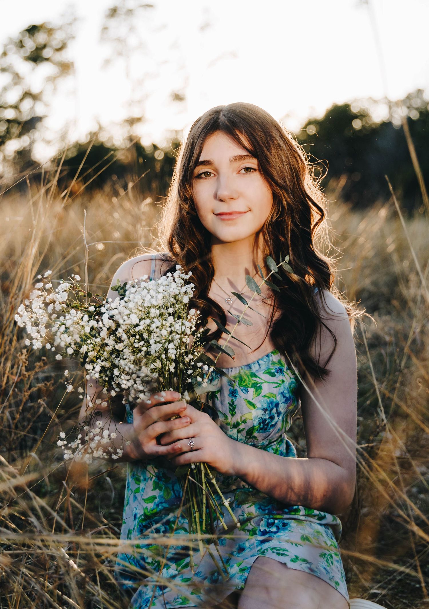 Young brunette woman holding white wildflowers in a floral dress in a sunlit field.