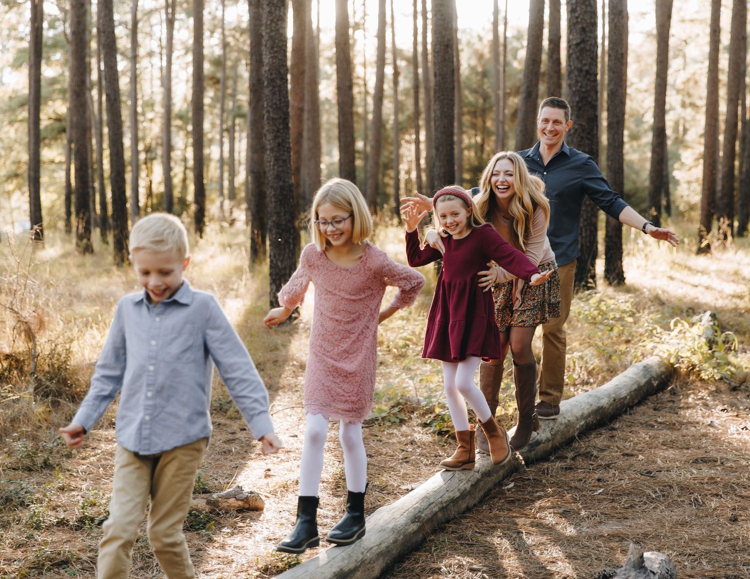 Joyful family of five playing on fallen log in sunny pine forest trail.