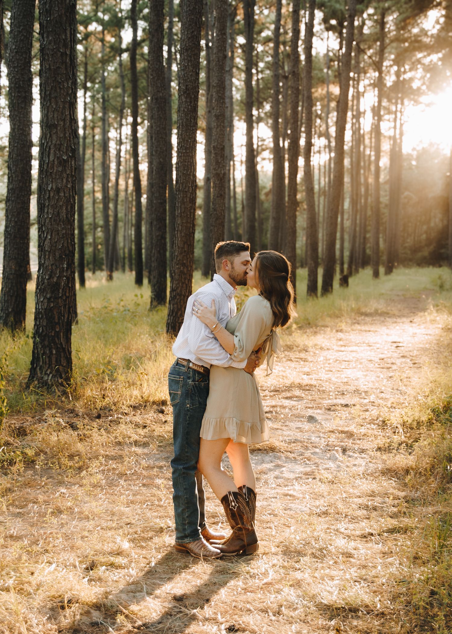 Couple embracing on a sunlit forest path surrounded by tall pine trees during golden hour.