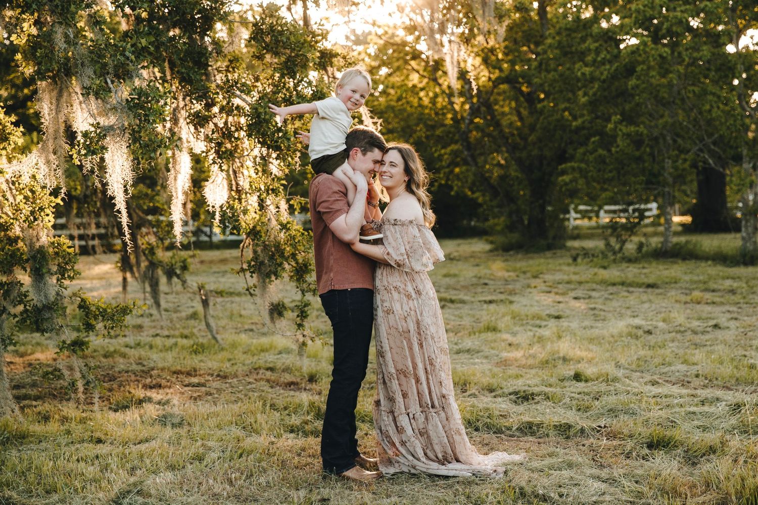 Family of three posing at golden hour in a lush green field, mother in elegant lace gown.