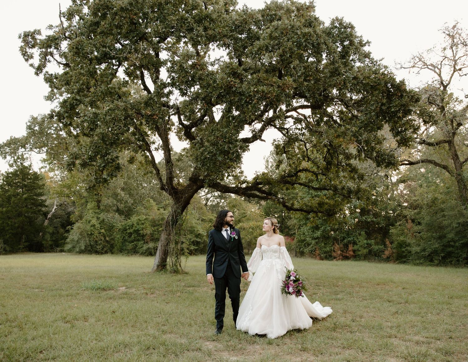 Bride and groom walking hand in hand across a green field with a large oak tree backdrop.