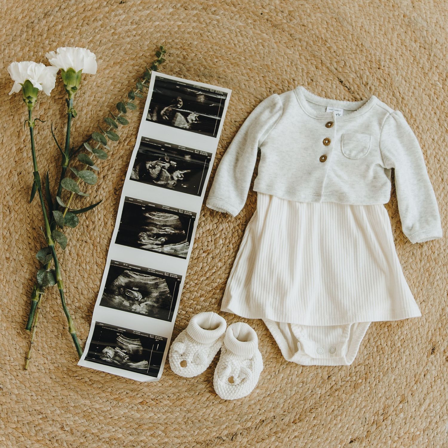 Flat lay of baby ultrasound photos, white floral outfit, booties, and eucalyptus on woven rug.
