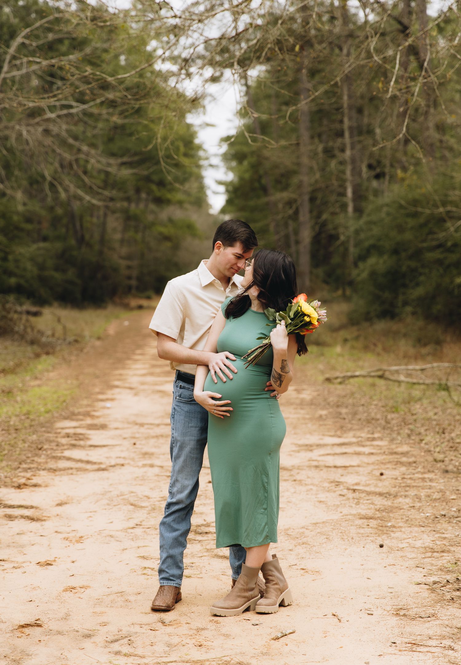 Couple embracing on forest dirt path during maternity session, woman in green dress holding sunflowers.