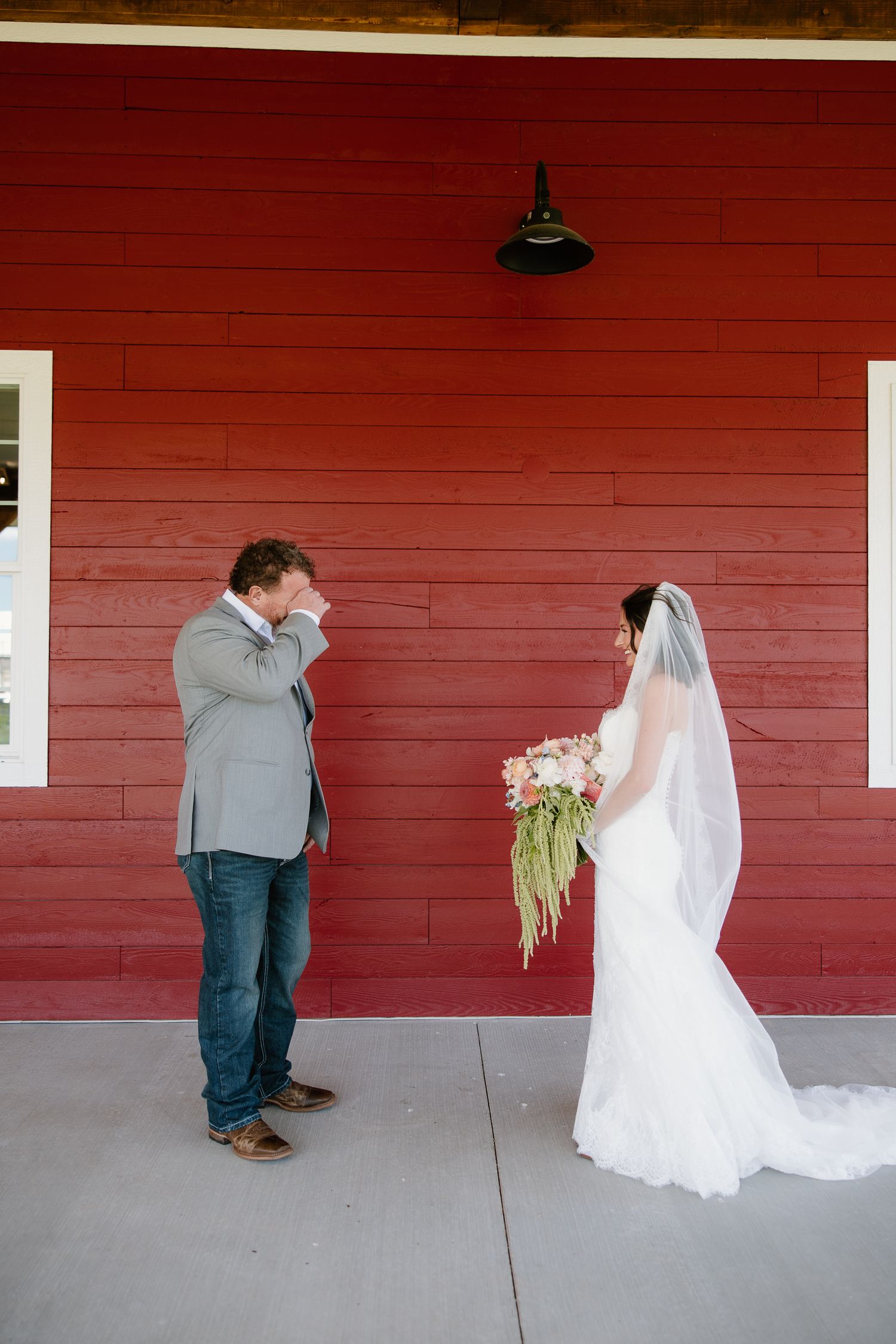 Groom wipes tears during emotional first look with bride in white gown holding bouquet outside red barn.
