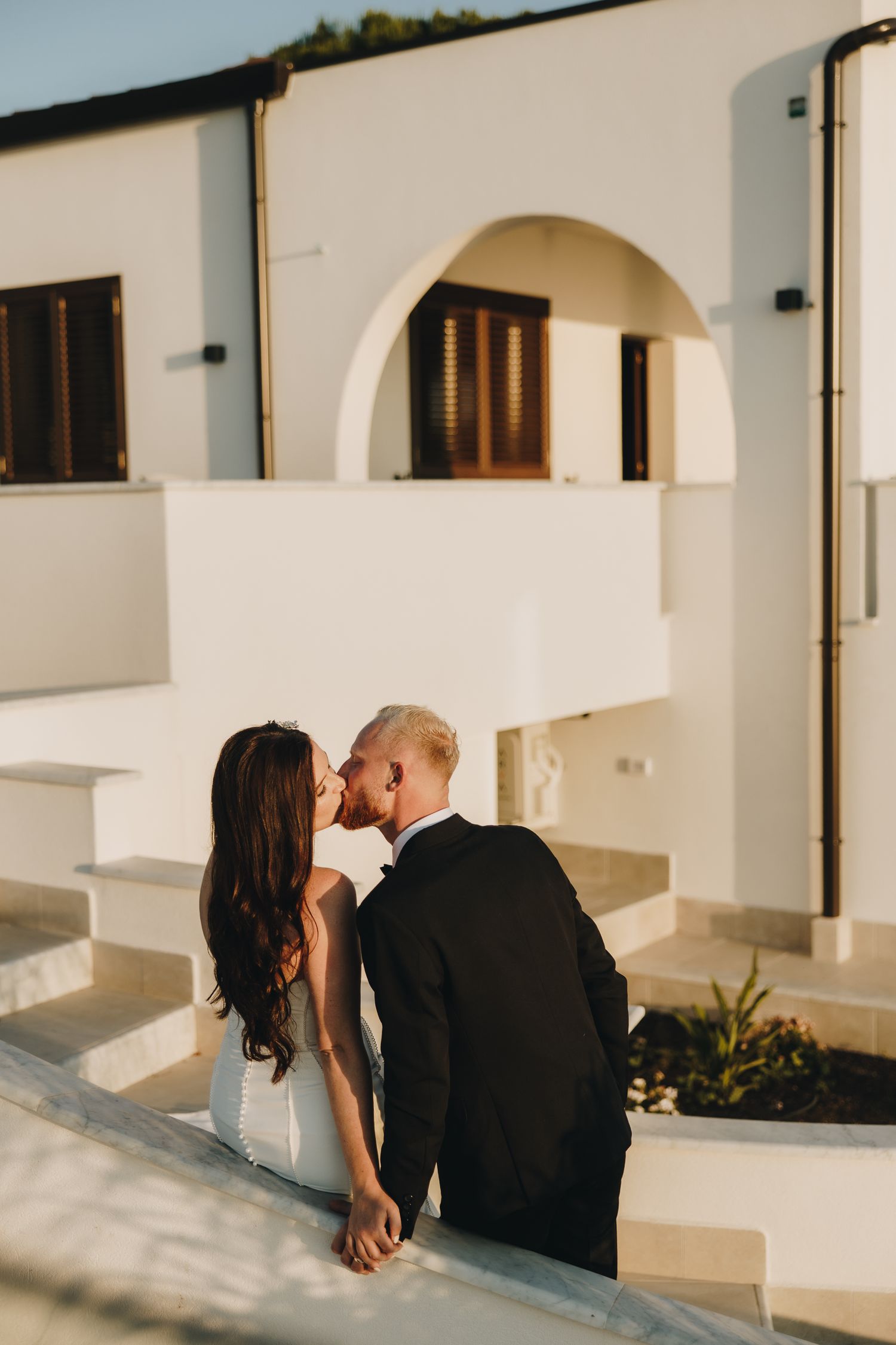 Bride and groom share a romantic kiss on white Mediterranean villa steps, holding hands in golden sunset light.