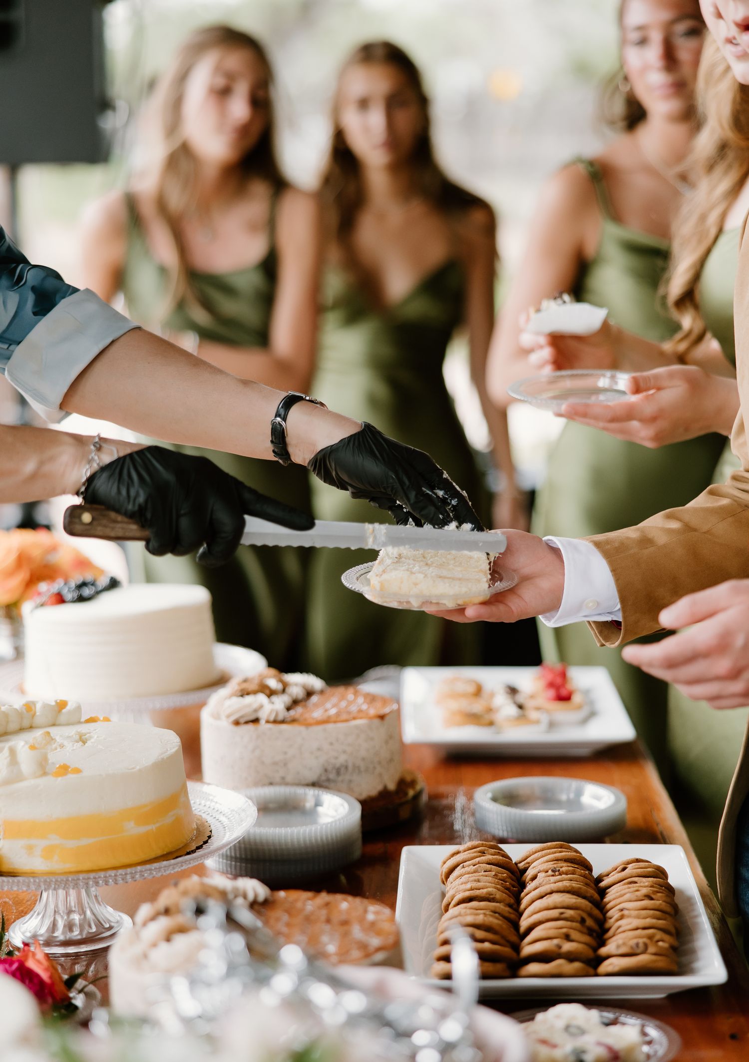 Catering staff in black gloves serve wedding cake at a dessert table with cookies and treats, as bridesmaids in sage green dresses look on.