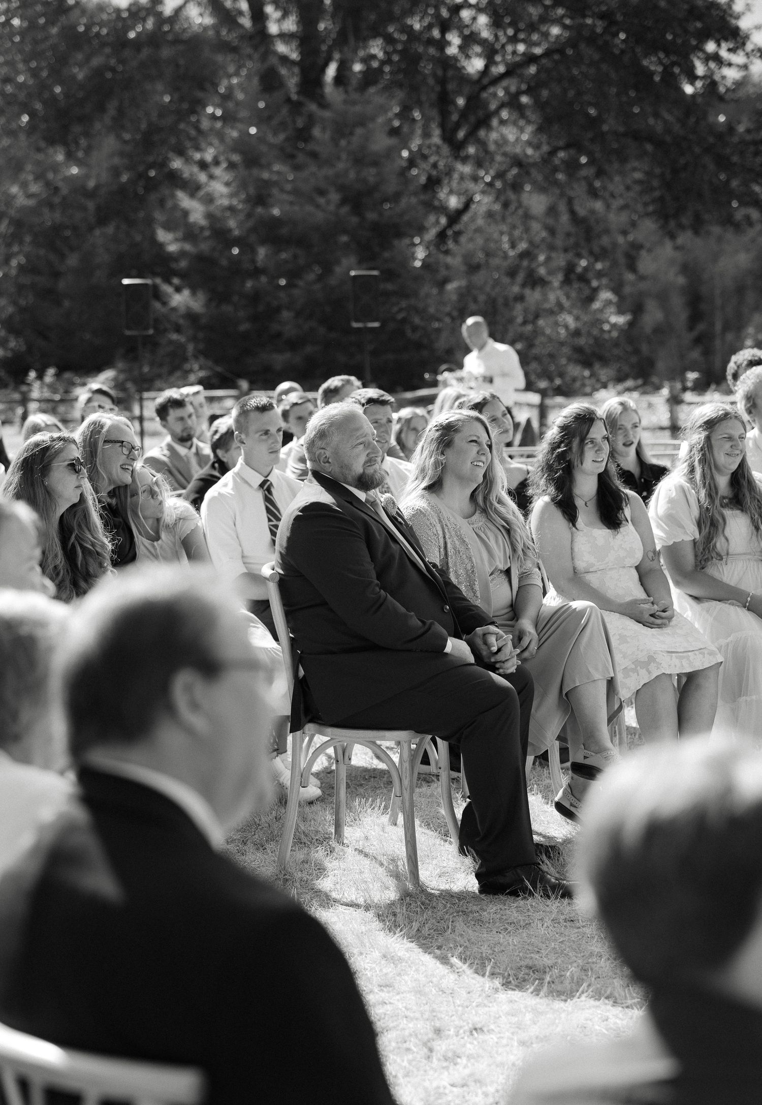 Wedding guests smile and laugh while seated at outdoor ceremony in black and white photo.