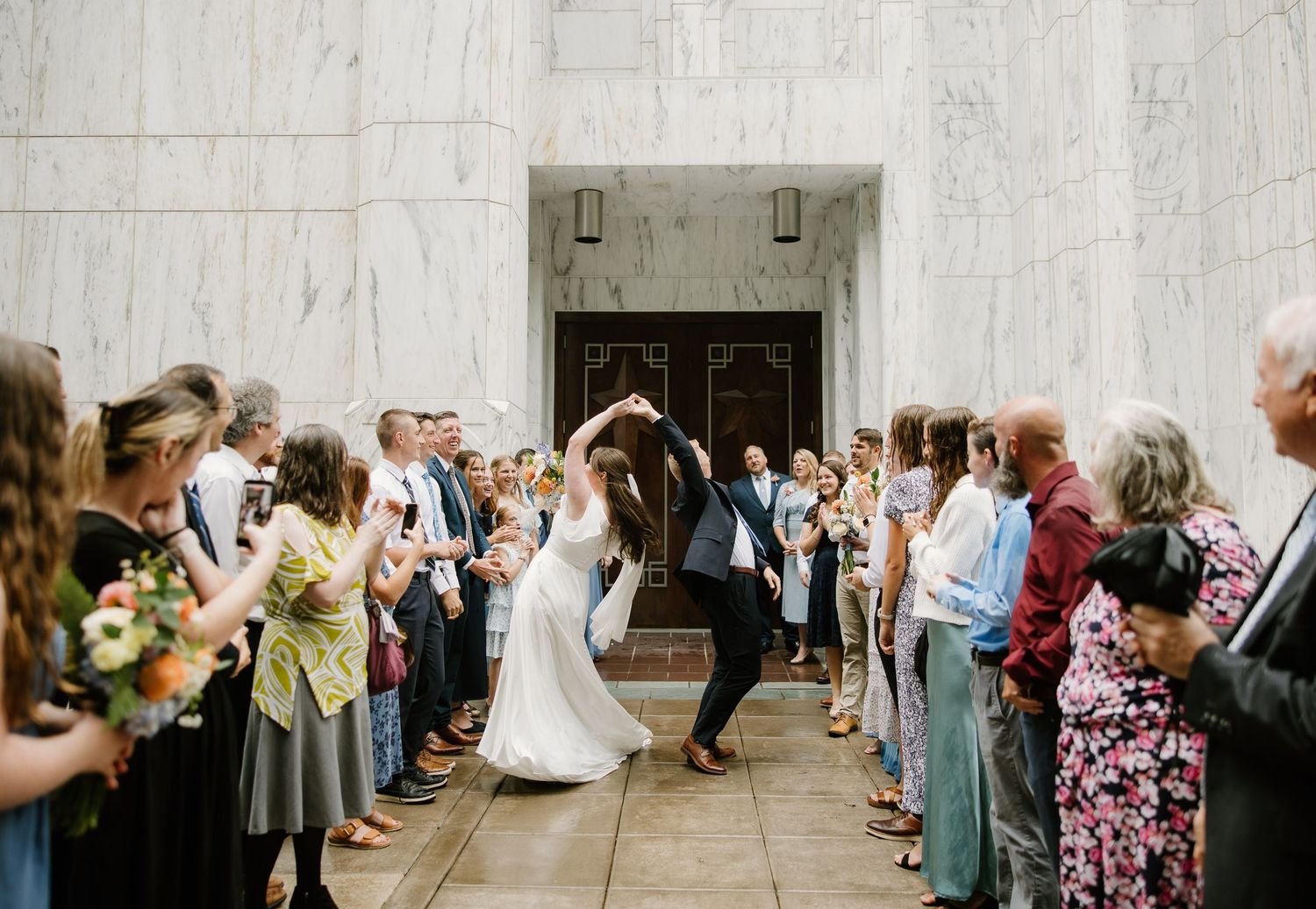 Bride and groom share a dance outside a grand marble building surrounded by cheering wedding guests.