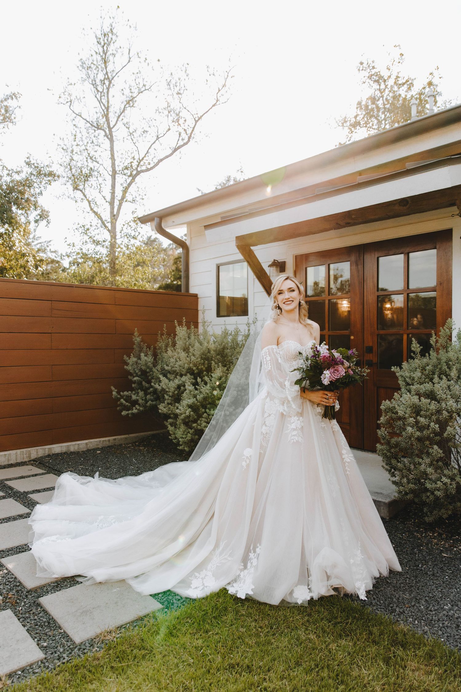 Radiant bride in a floral ballgown with long veil poses outdoors holding a bouquet in golden light.