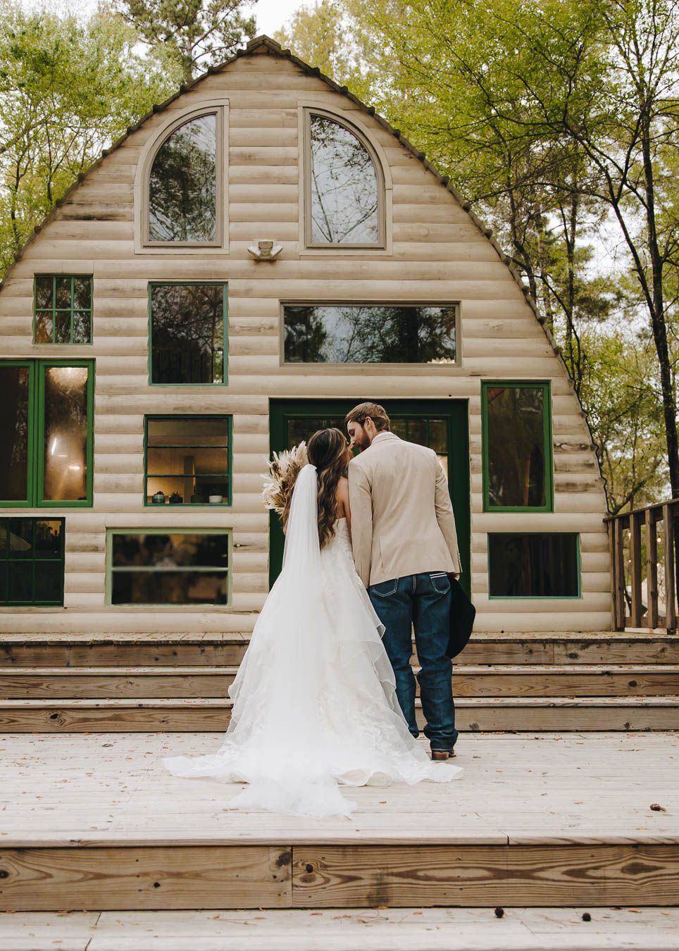 Bride and groom stand together facing a rustic log cabin wedding venue surrounded by lush green trees.