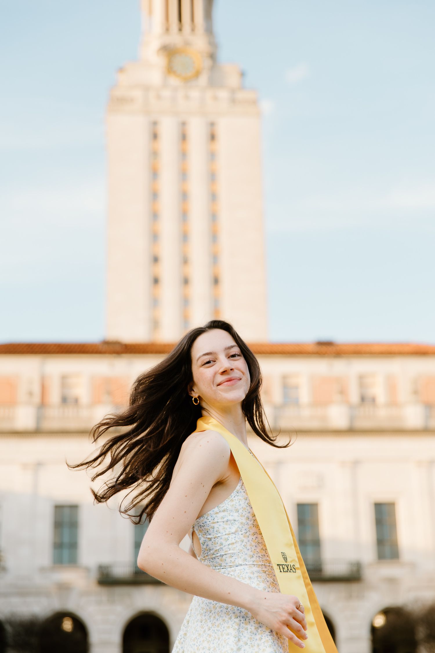 Brunette woman in a white dress wearing a UT Texas graduation sash posing in front of the UT Tower.