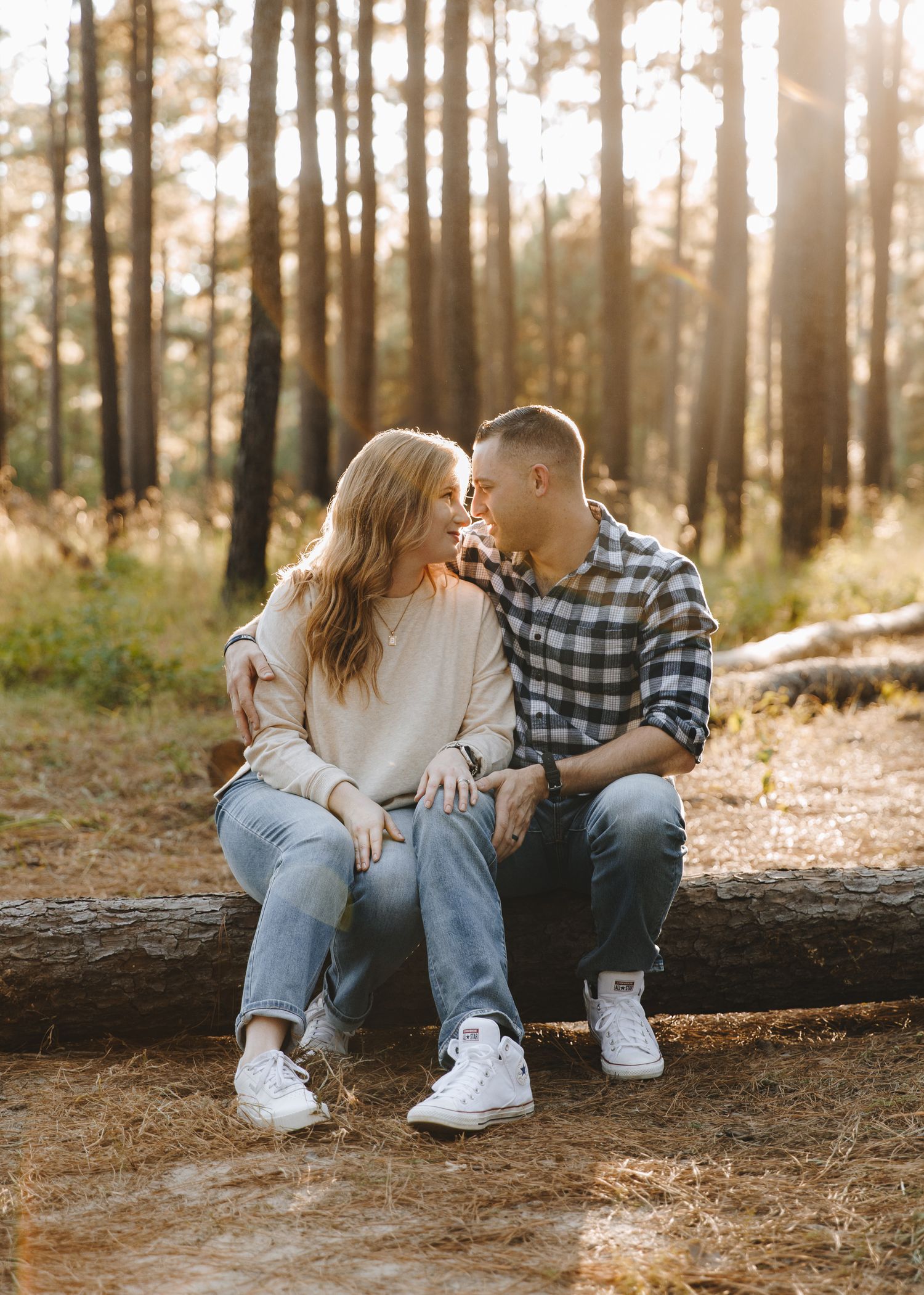 A couple sits closely on a fallen log in a golden sunlit forest, sharing a romantic moment.