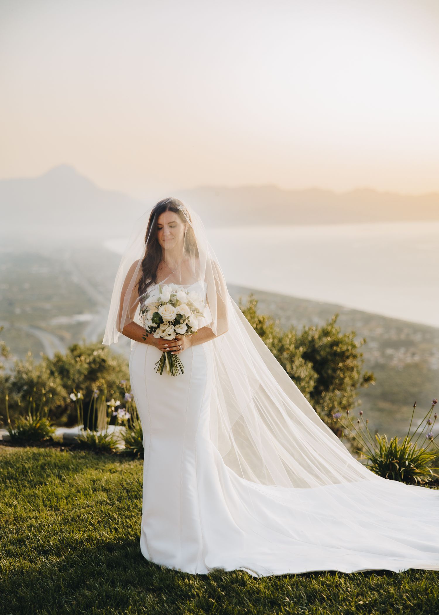 Bride in white gown and veil holds bouquet at sunset with coastal mountain views behind her.