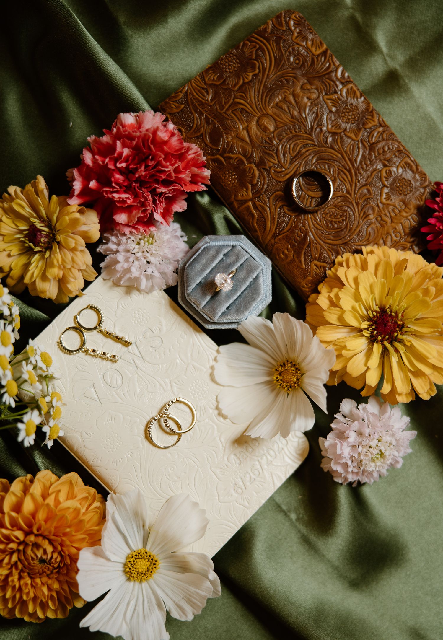 Wedding flat lay with rings, keys, flowers, and a velvet ring box on green silk.