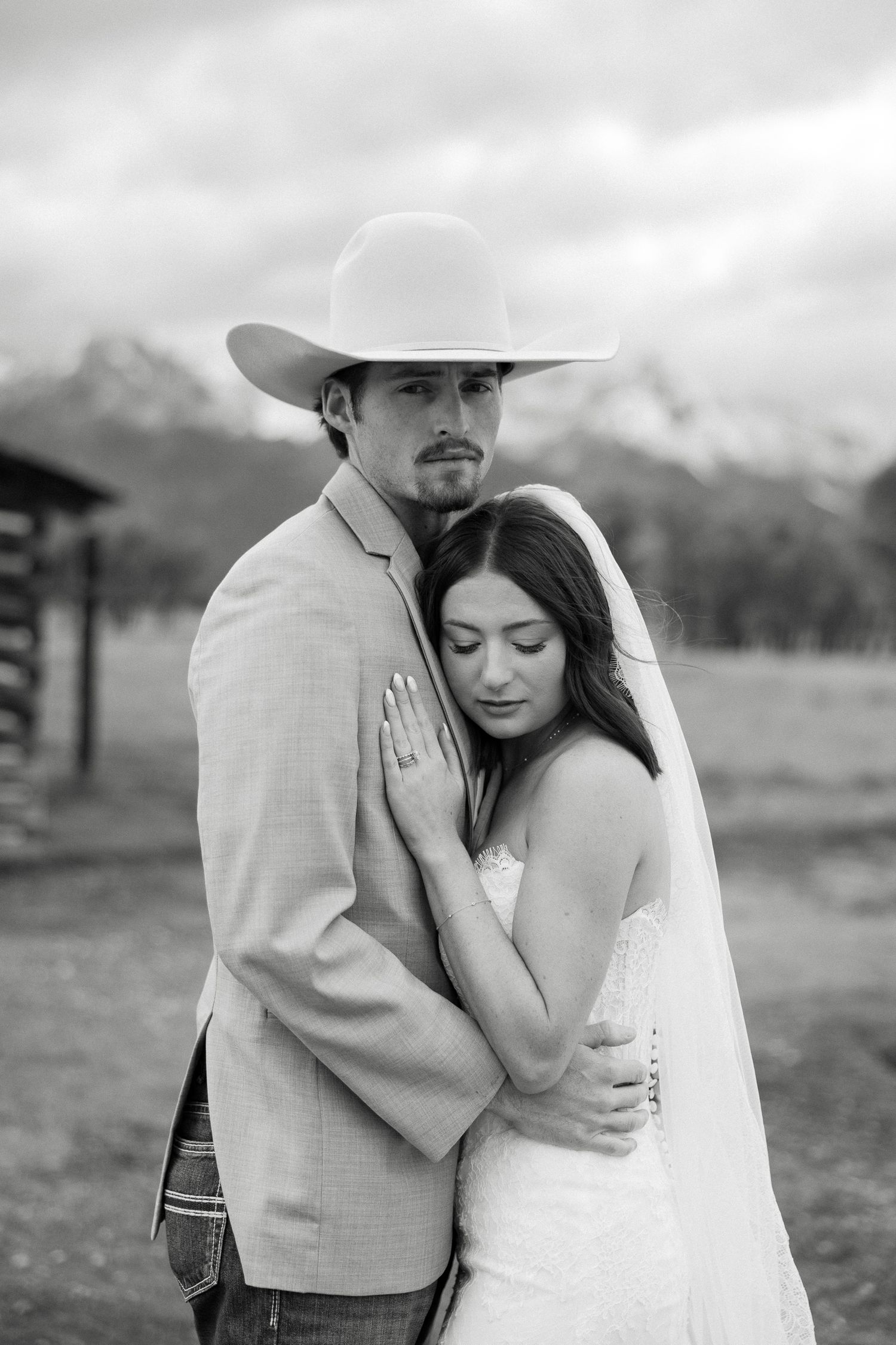 Black and white photo of a bride and groom embracing outdoors, groom wearing a cowboy hat with mountains behind.