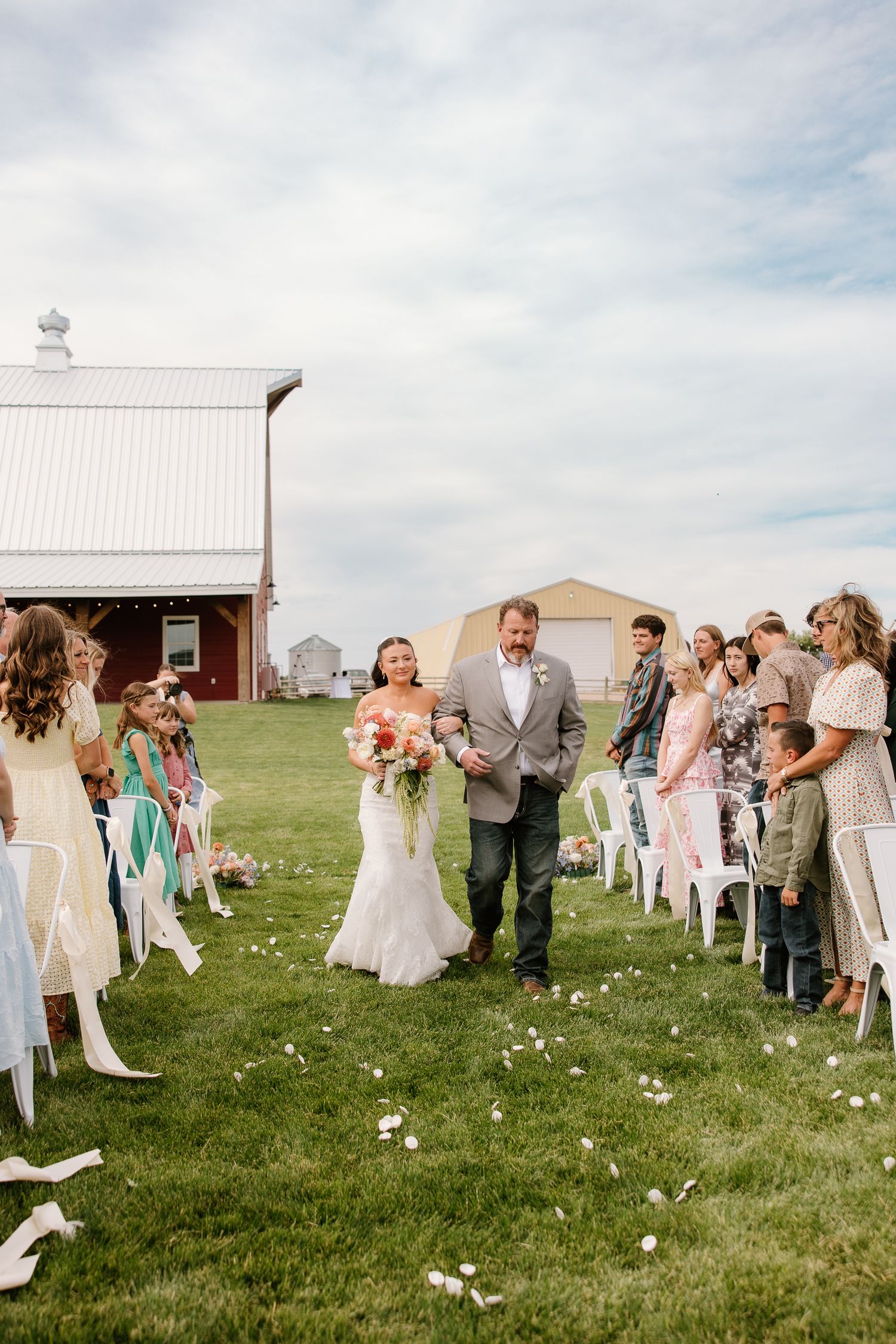 Bride walking down outdoor aisle with escort past seated guests at farm wedding.