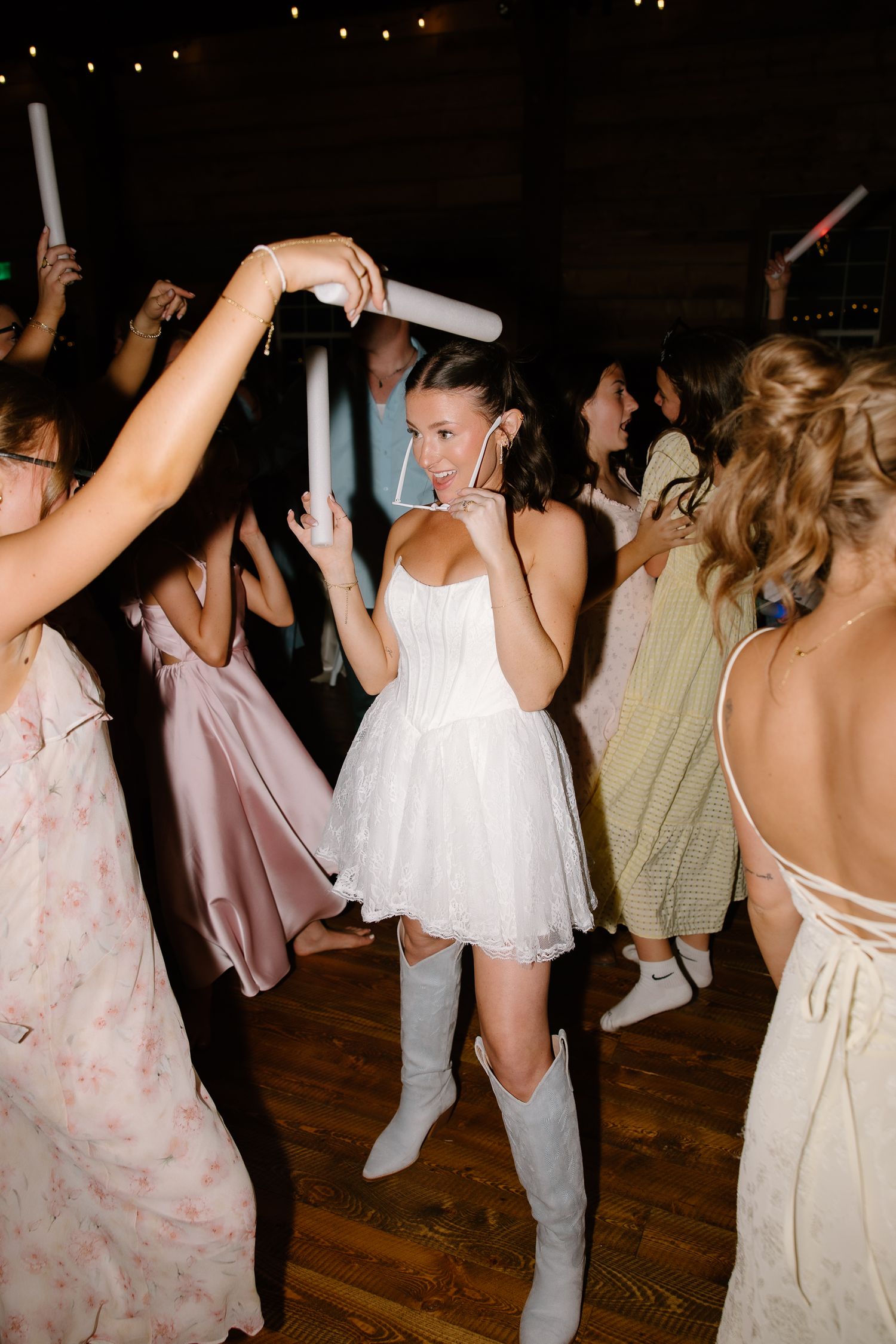 Bride in short white dress and gray cowboy boots dancing with guests holding glow sticks at wedding reception.