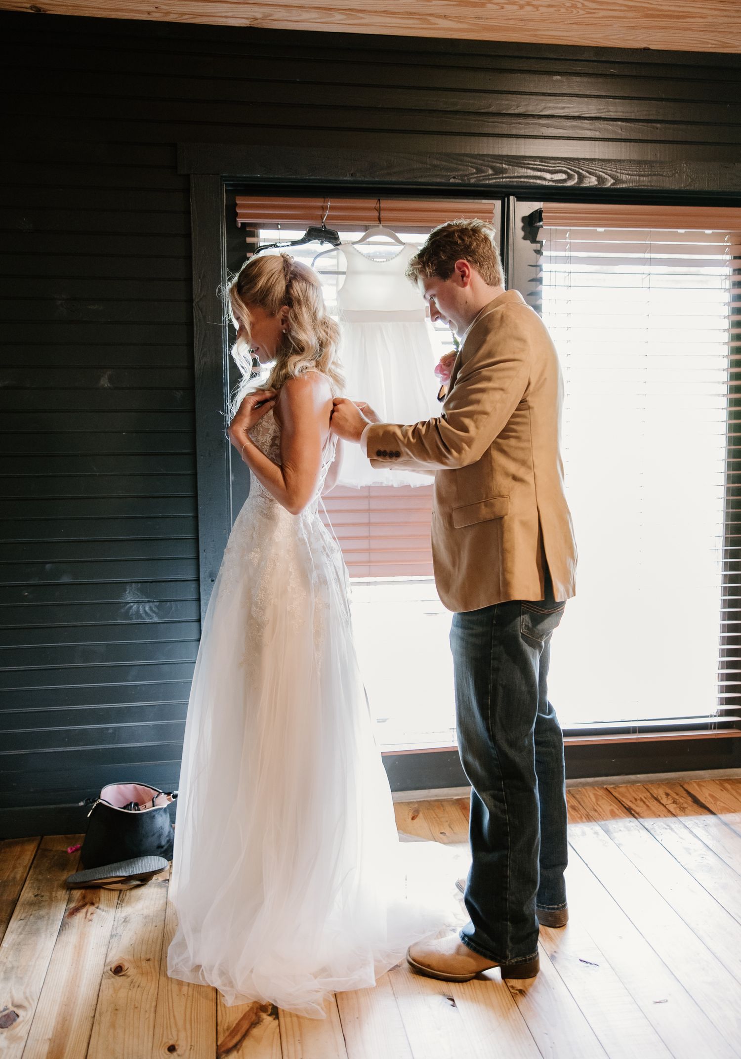 Groom in tan blazer fastening bride's lace wedding dress near window in rustic cabin.
