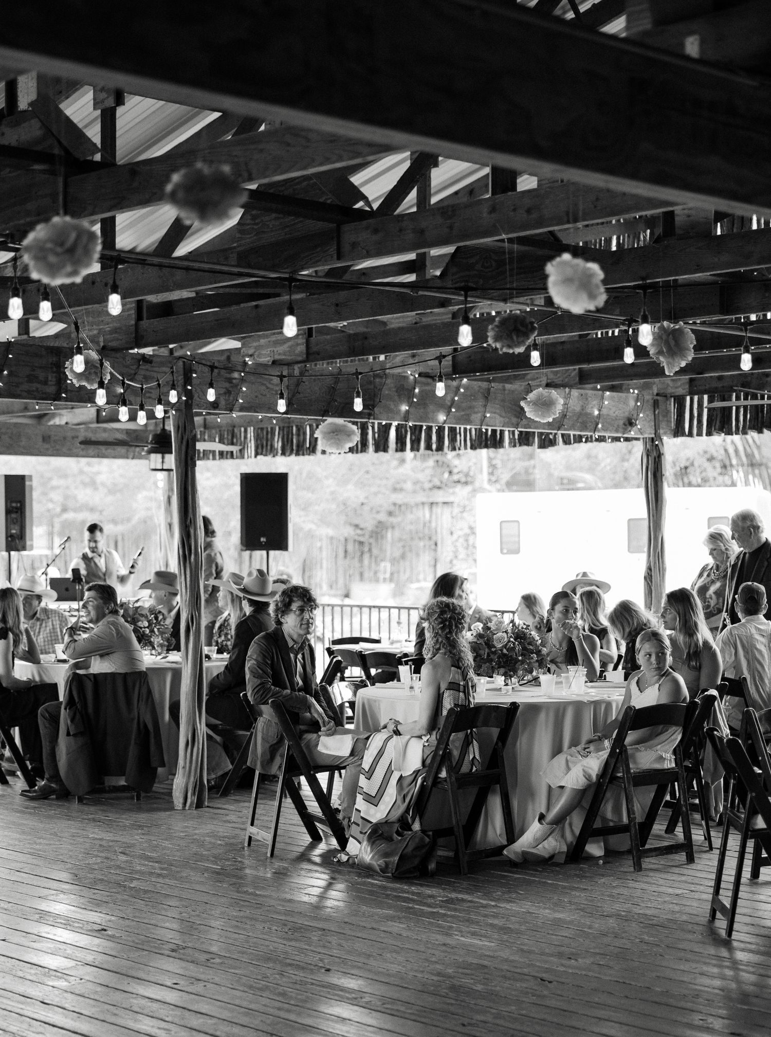 Black and white photo of wedding reception guests dining under string lights in rustic open-air venue.
