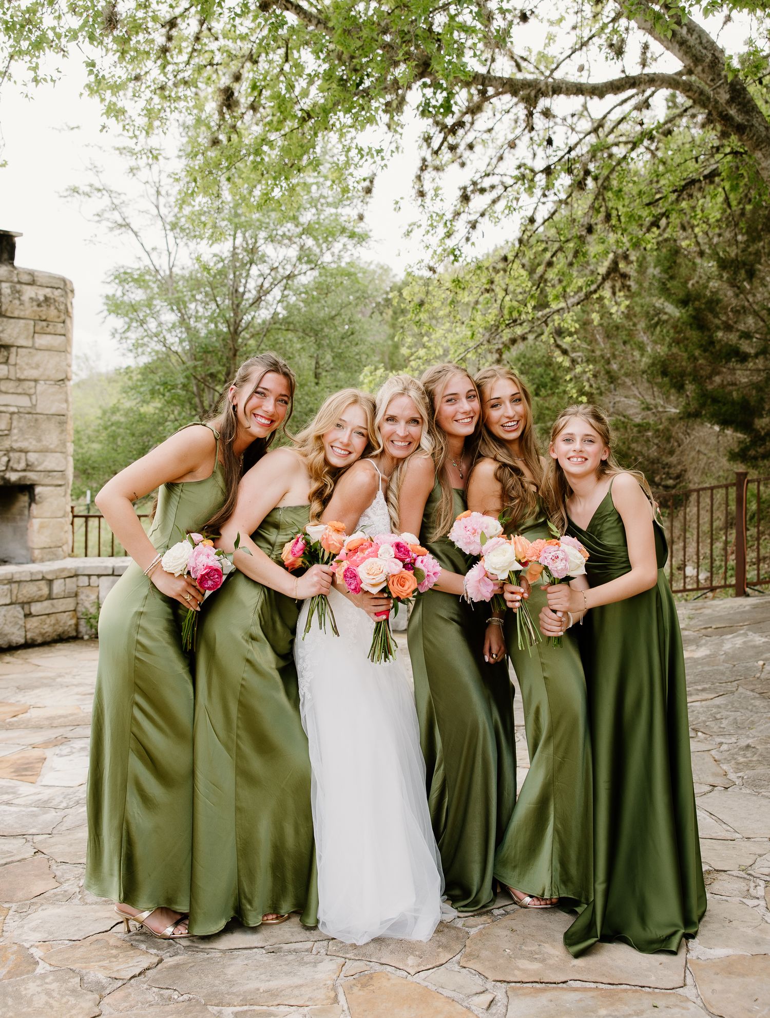 Bride poses with five bridesmaids wearing olive green satin dresses holding colorful bouquets outdoors.