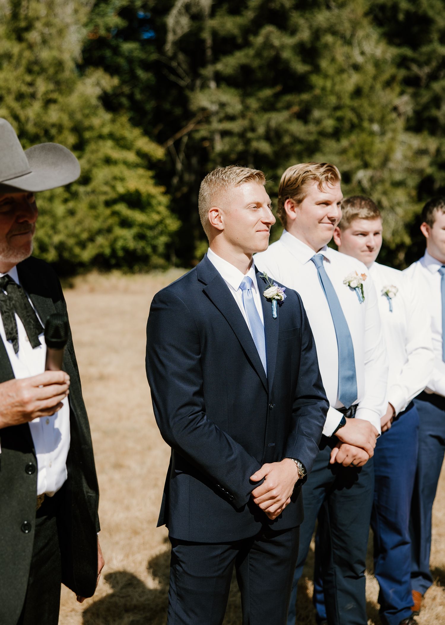 Groom and groomsmen in navy suits stand outdoors at a sunny wedding ceremony.
