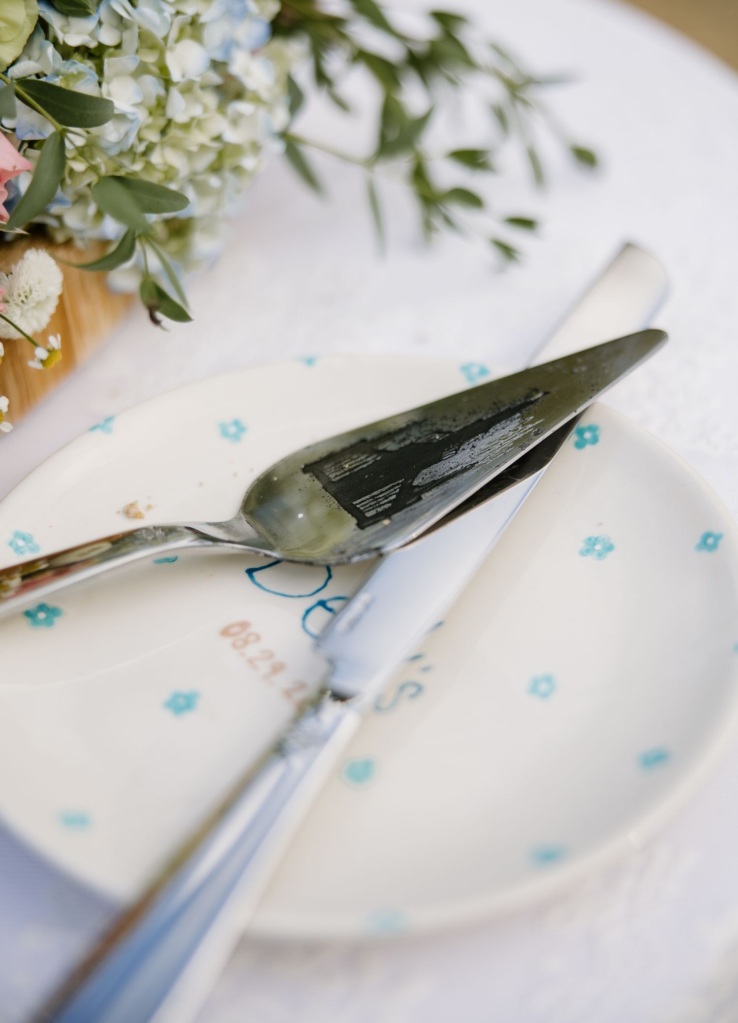 Decorated wedding cake plate with silver serving utensils and blue floral details on white tablecloth.