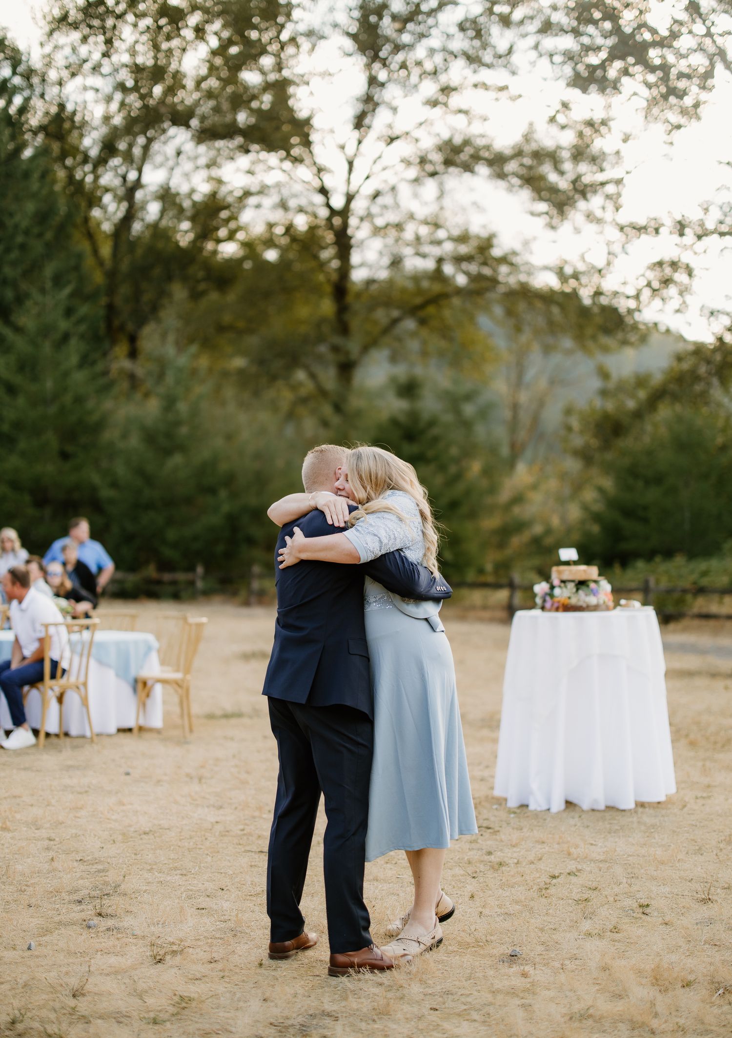 Couple shares emotional dance at outdoor wedding reception with cake table in background.