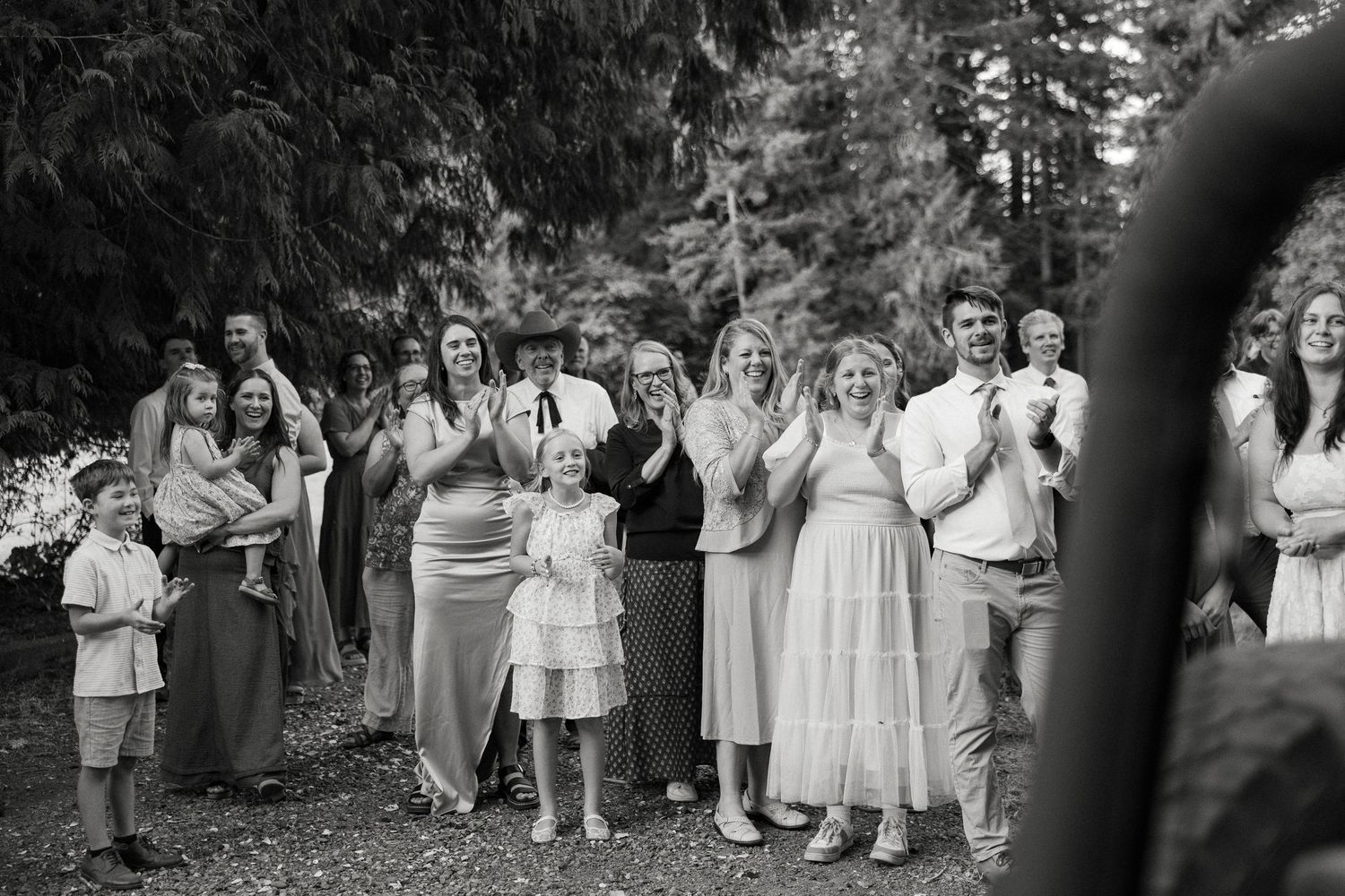 Black and white photo of joyful wedding guests clapping and celebrating outdoors among tall trees.