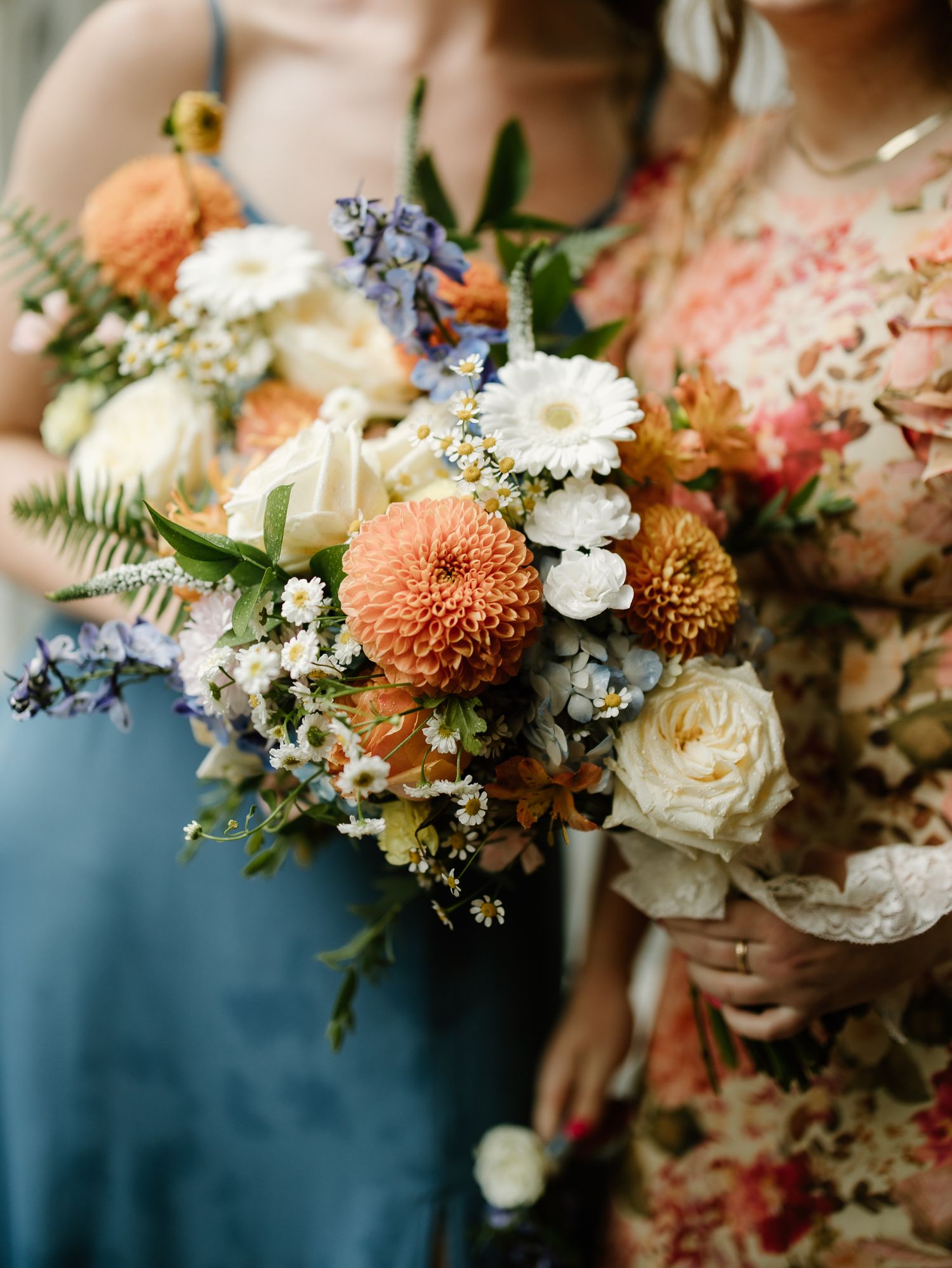 Bridesmaids hold lush bouquets of orange dahlias, white roses, and blue wildflowers at a wedding.