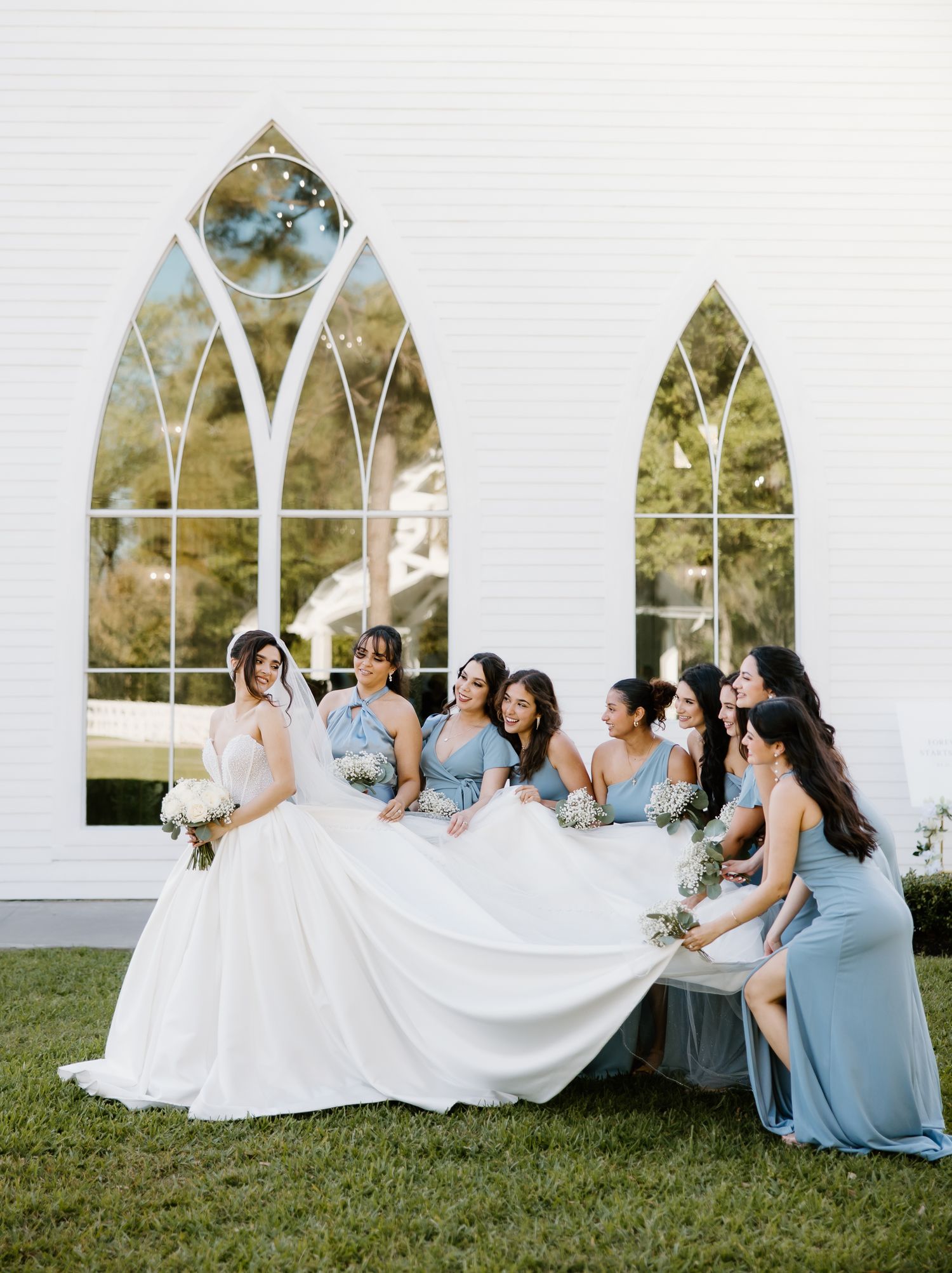 Bride in ballgown surrounded by bridesmaids in dusty blue dresses before white chapel windows.