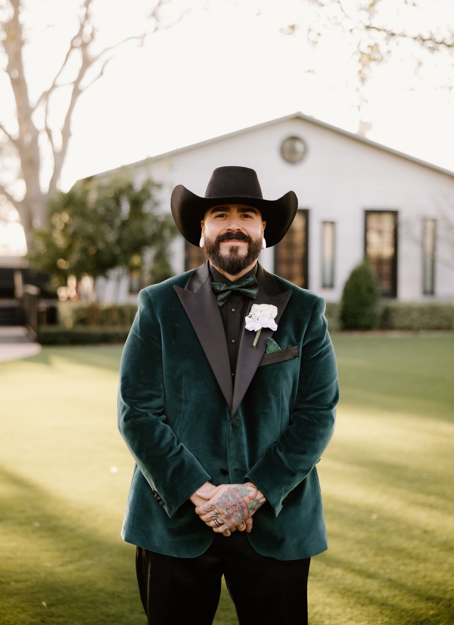 Groom in green velvet tuxedo and black cowboy hat with white boutonniere outdoors.