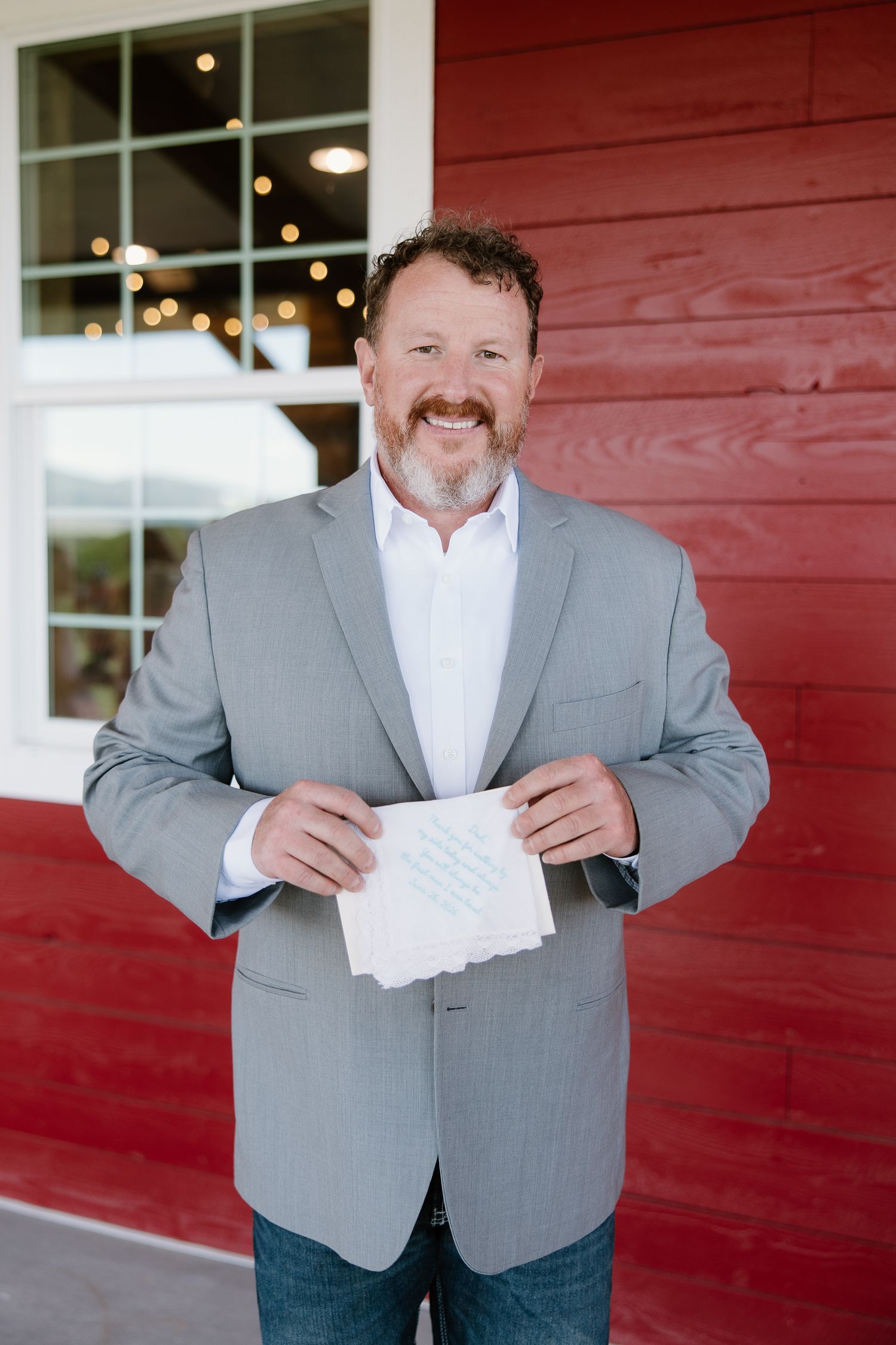 Smiling man in gray blazer holds a handwritten note outside a red barn.