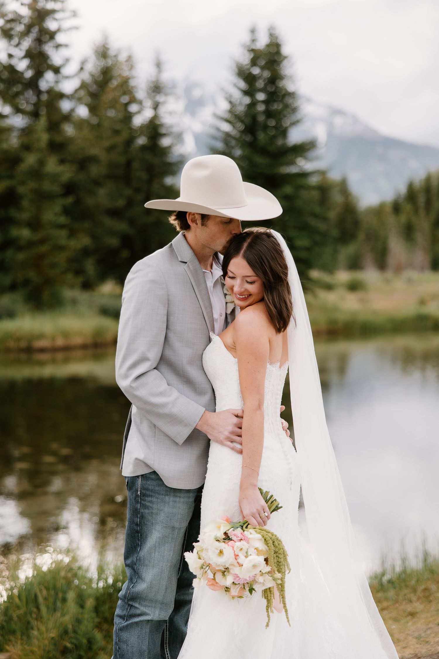 Bride and groom sharing a romantic moment by a mountain lake, groom in cowboy hat, bride holding a floral bouquet.