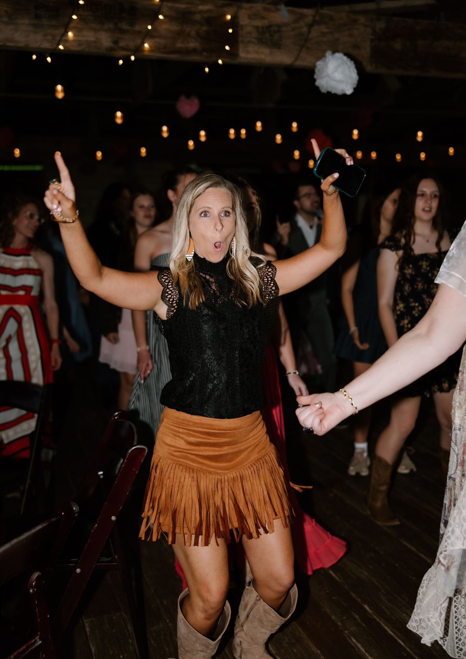 Woman in a fringed suede skirt and cowboy boots dancing energetically at a lively wedding reception.