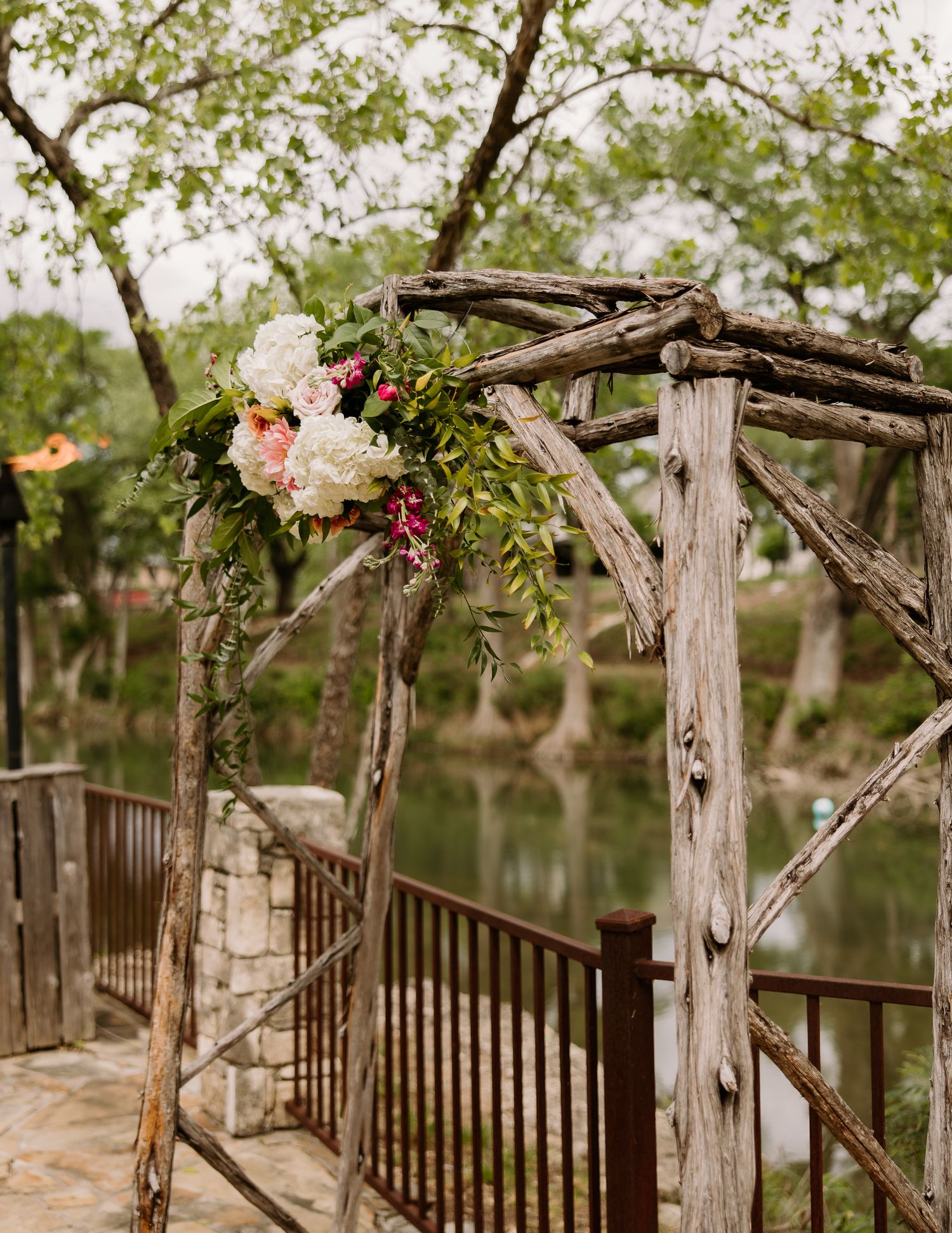 Rustic wooden wedding arch adorned with pink and white floral arrangements overlooking a serene pond.