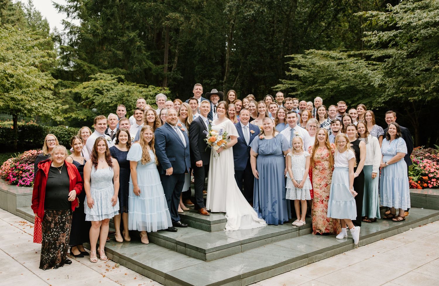 Large wedding party group photo with bride and groom surrounded by guests on steps in a lush garden setting.