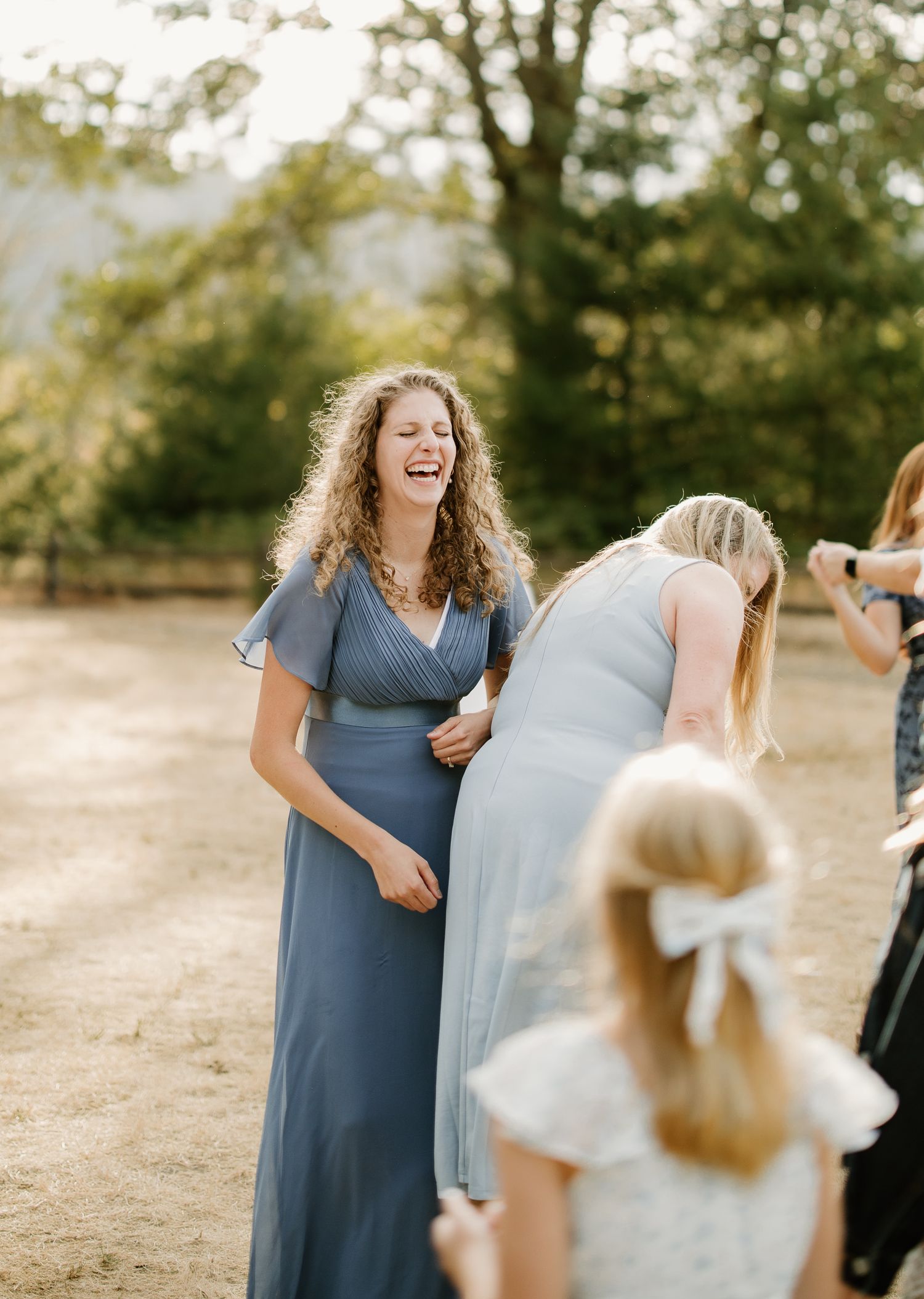 Bridesmaid in dusty blue dress laughing joyfully at outdoor wedding reception in golden light.