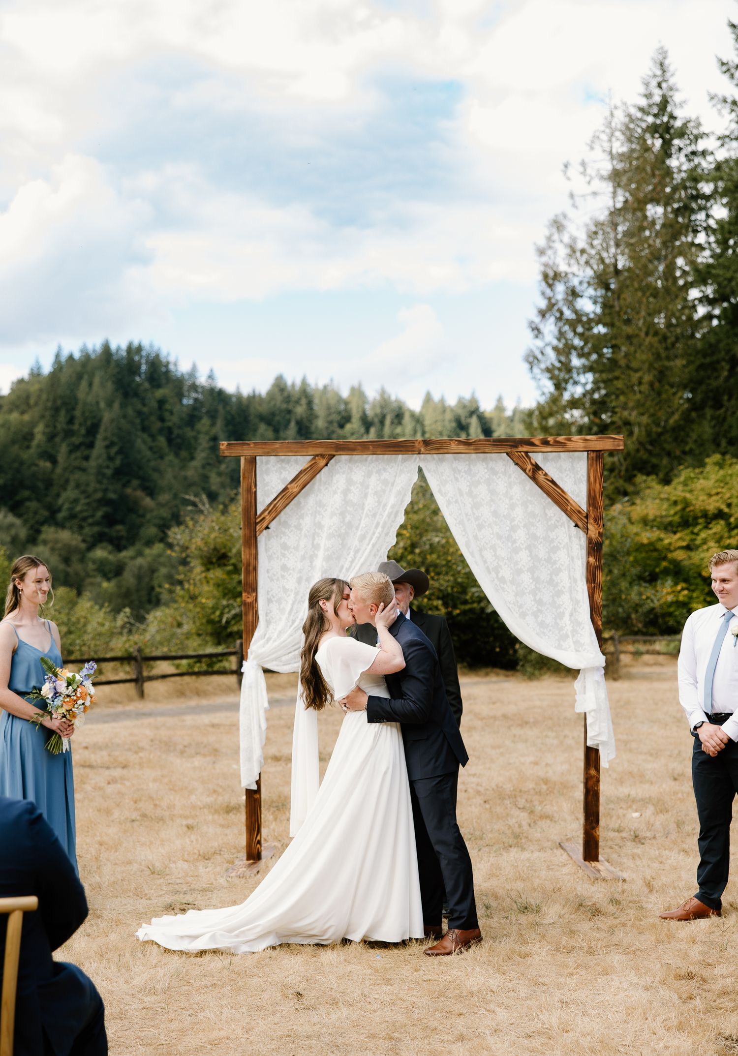 Bride and groom share first kiss under rustic wooden arch at outdoor meadow wedding.