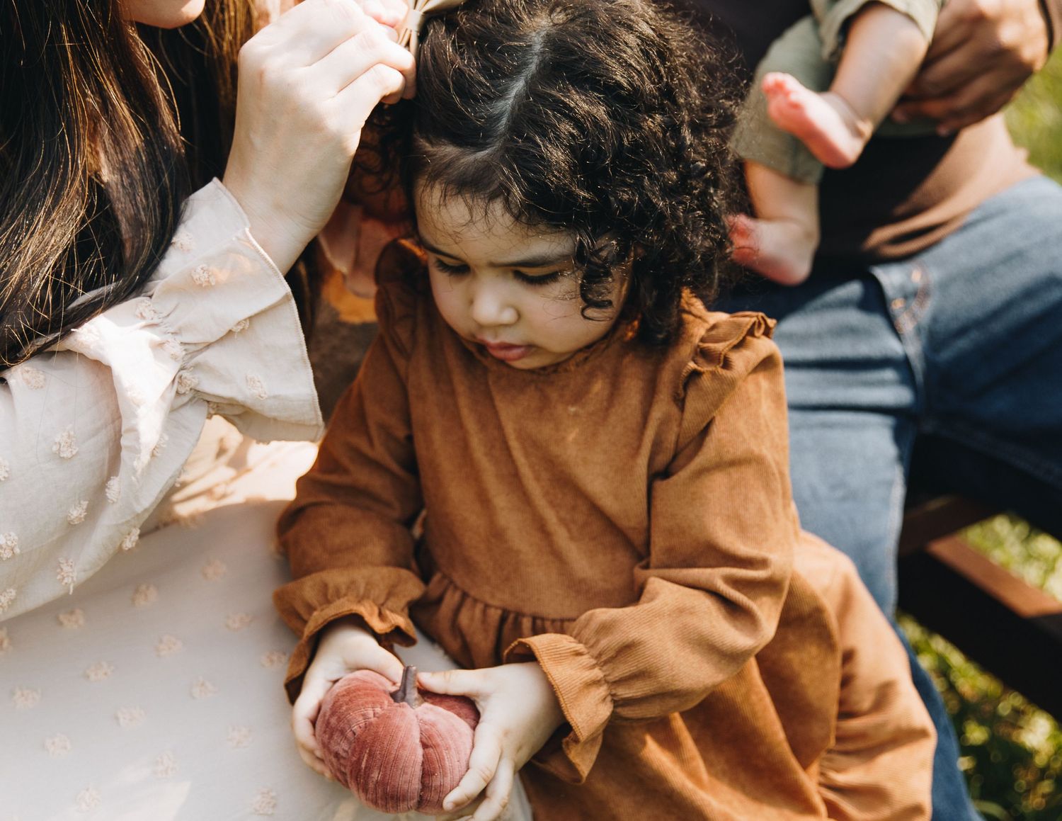 Young curly-haired toddler in brown dress holds a peach while sitting with family outdoors.