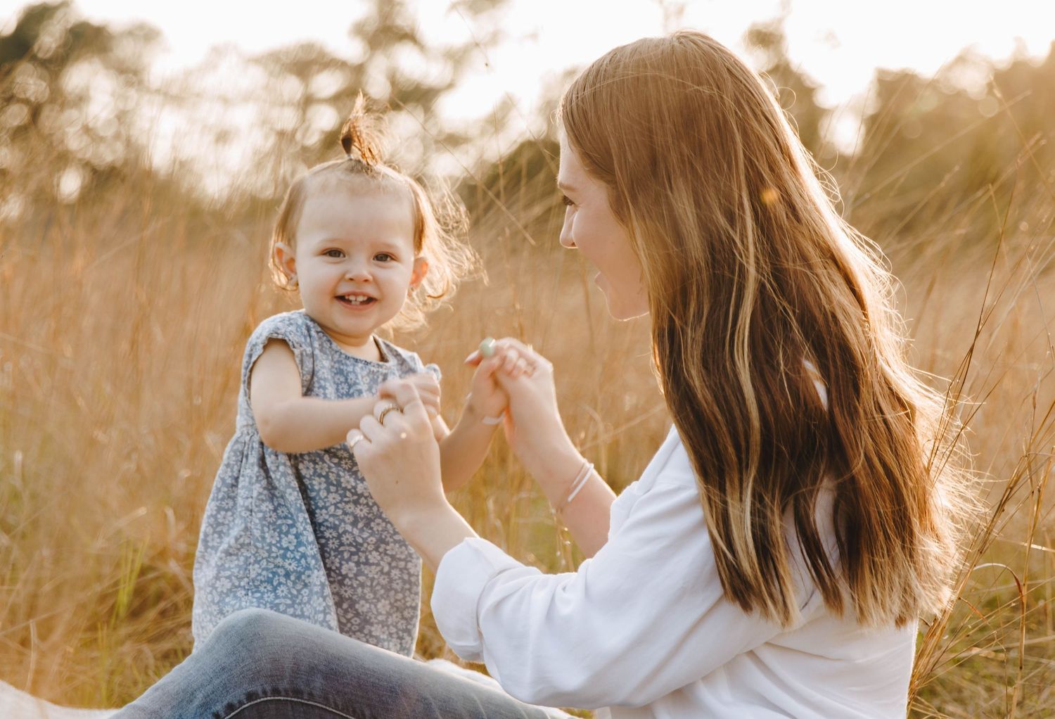 Mother plays with her smiling baby girl in a golden sunlit field of tall grass.